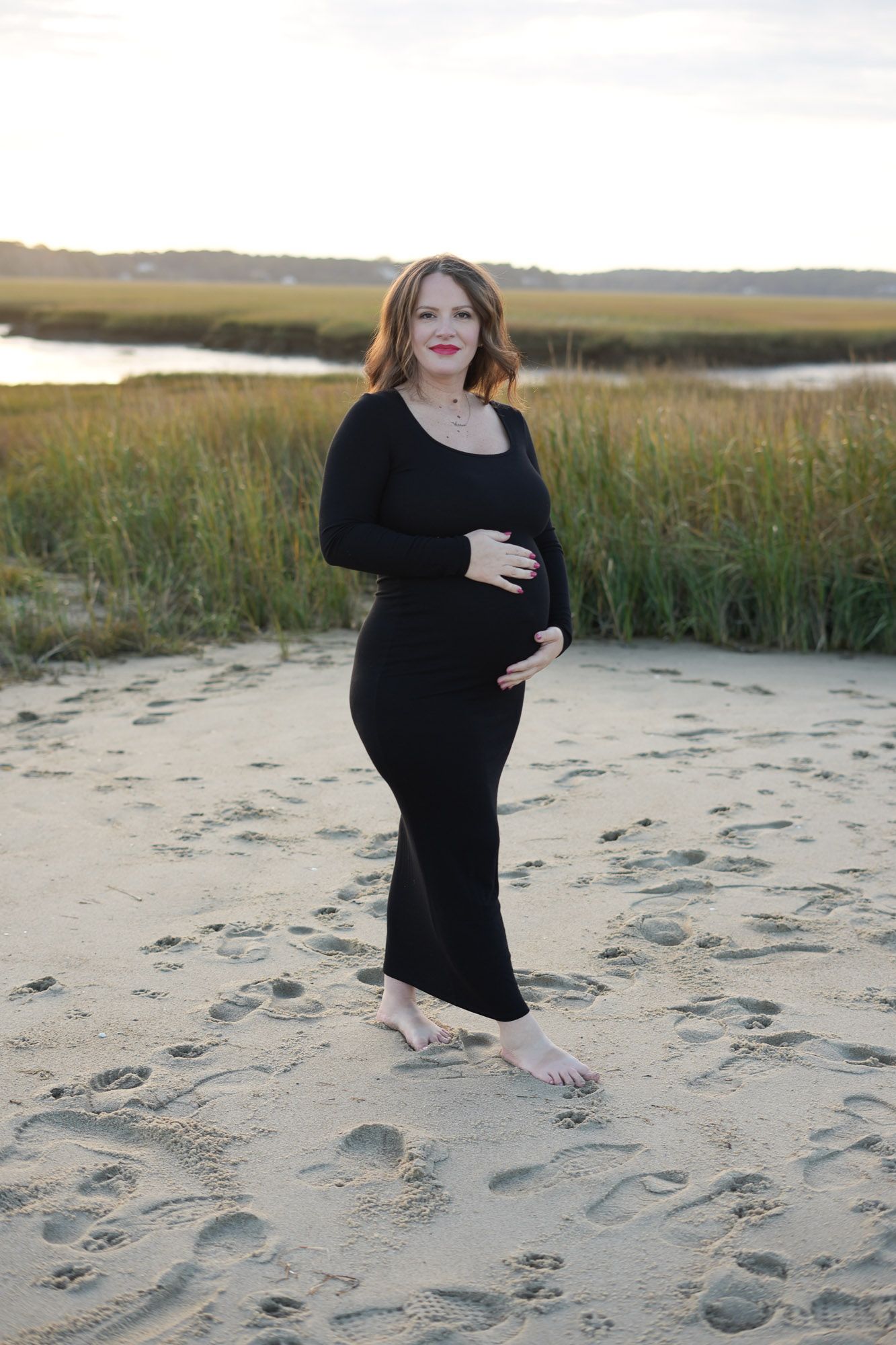 Barefoot pregnant woman in black dress posing on sandy beach with marsh background.