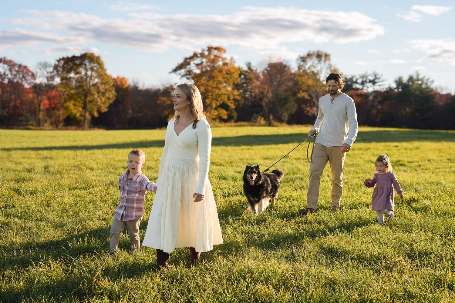 Family with pregnant mother, two children, and black dog walking in green autumn field.