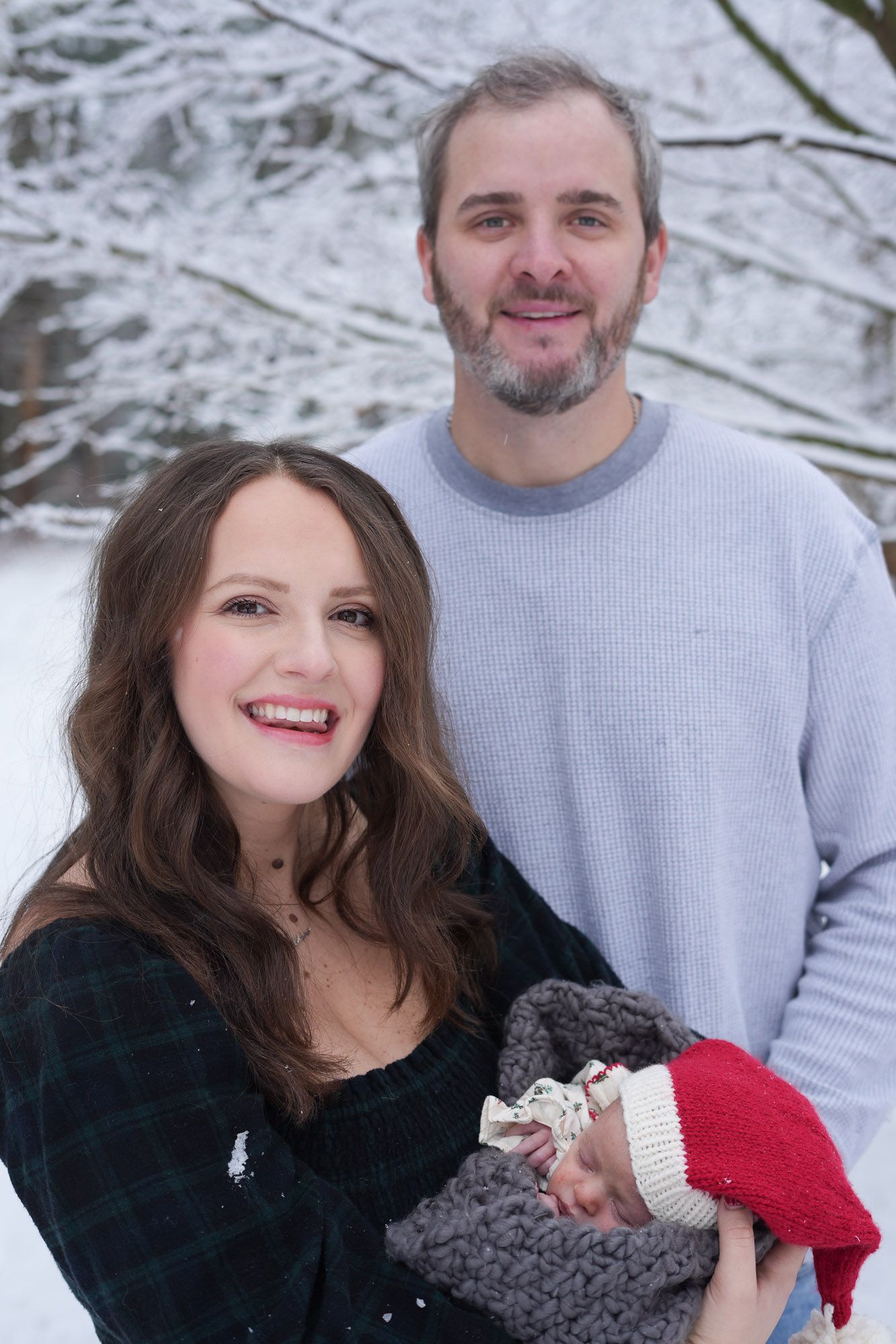 Happy couple smiling outdoors in snow holding newborn baby wearing red Santa hat in winter.