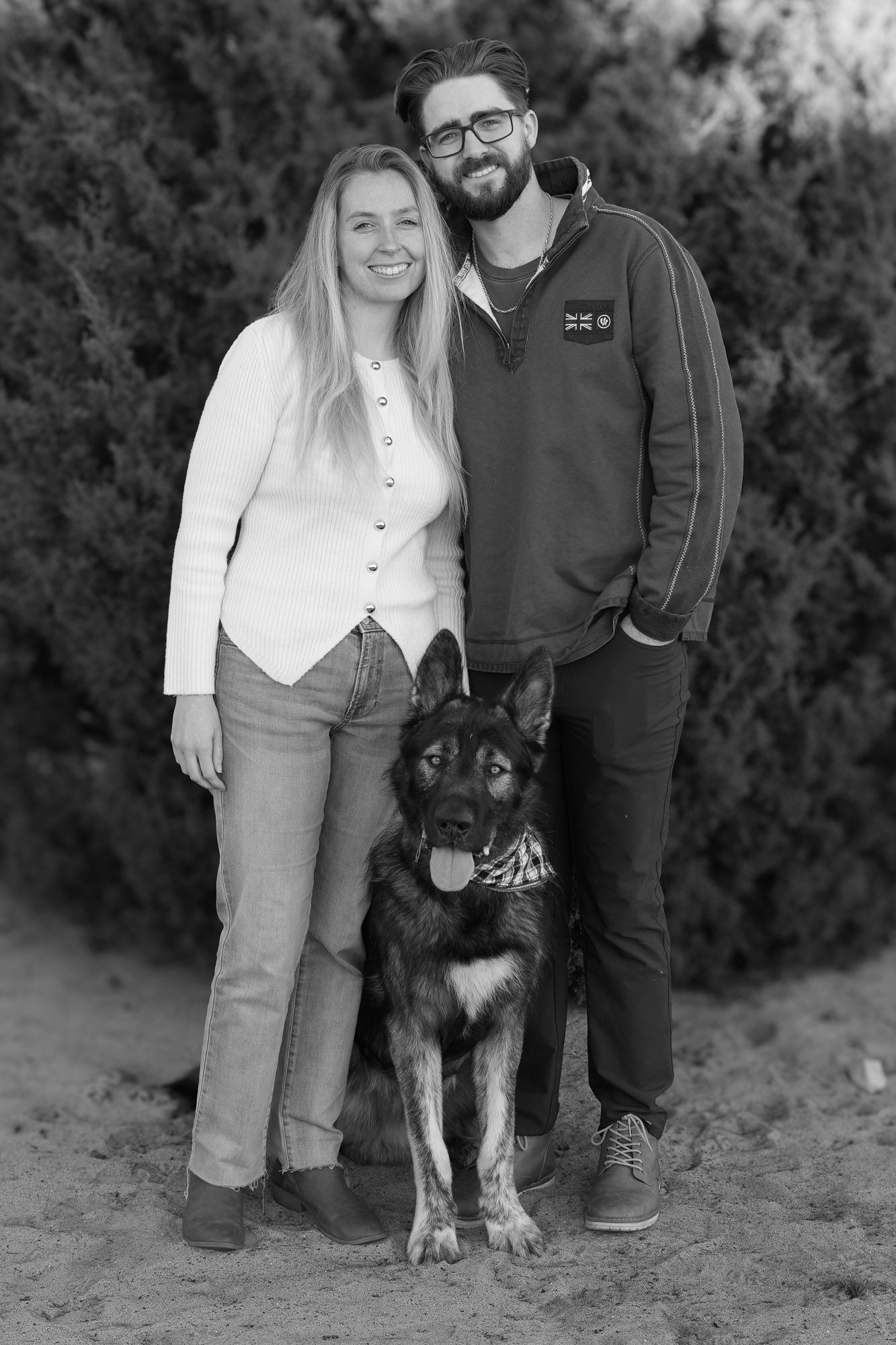 Black and white portrait of a smiling couple posing with their German Shepherd dog outdoors.