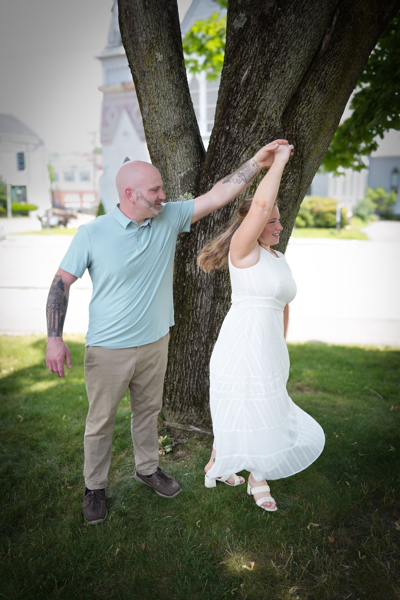 A bald man spins a woman in a white dress near a large tree on a sunny day.