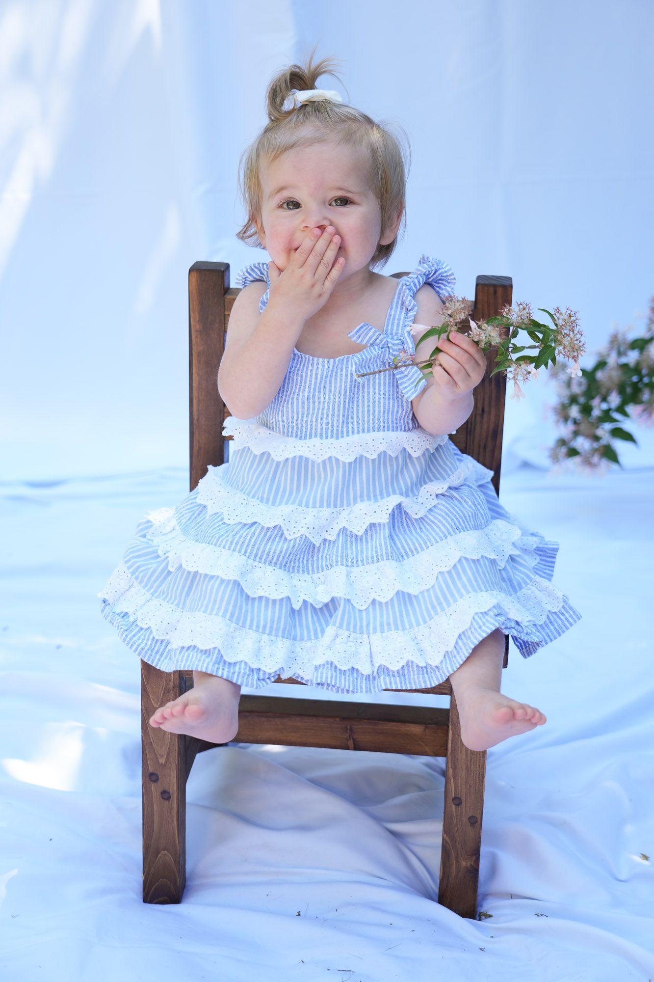 Toddler girl in blue ruffled dress sitting on wooden chair holding flowers with hand over mouth.
