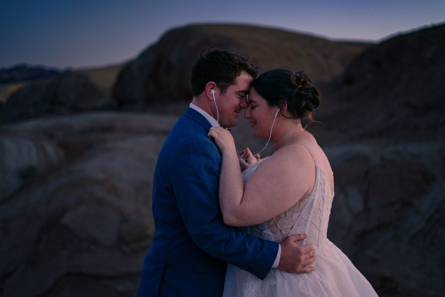 Newlyweds embracing tenderly at dusk on rocky terrain, sharing earbuds during their desert elopement.
