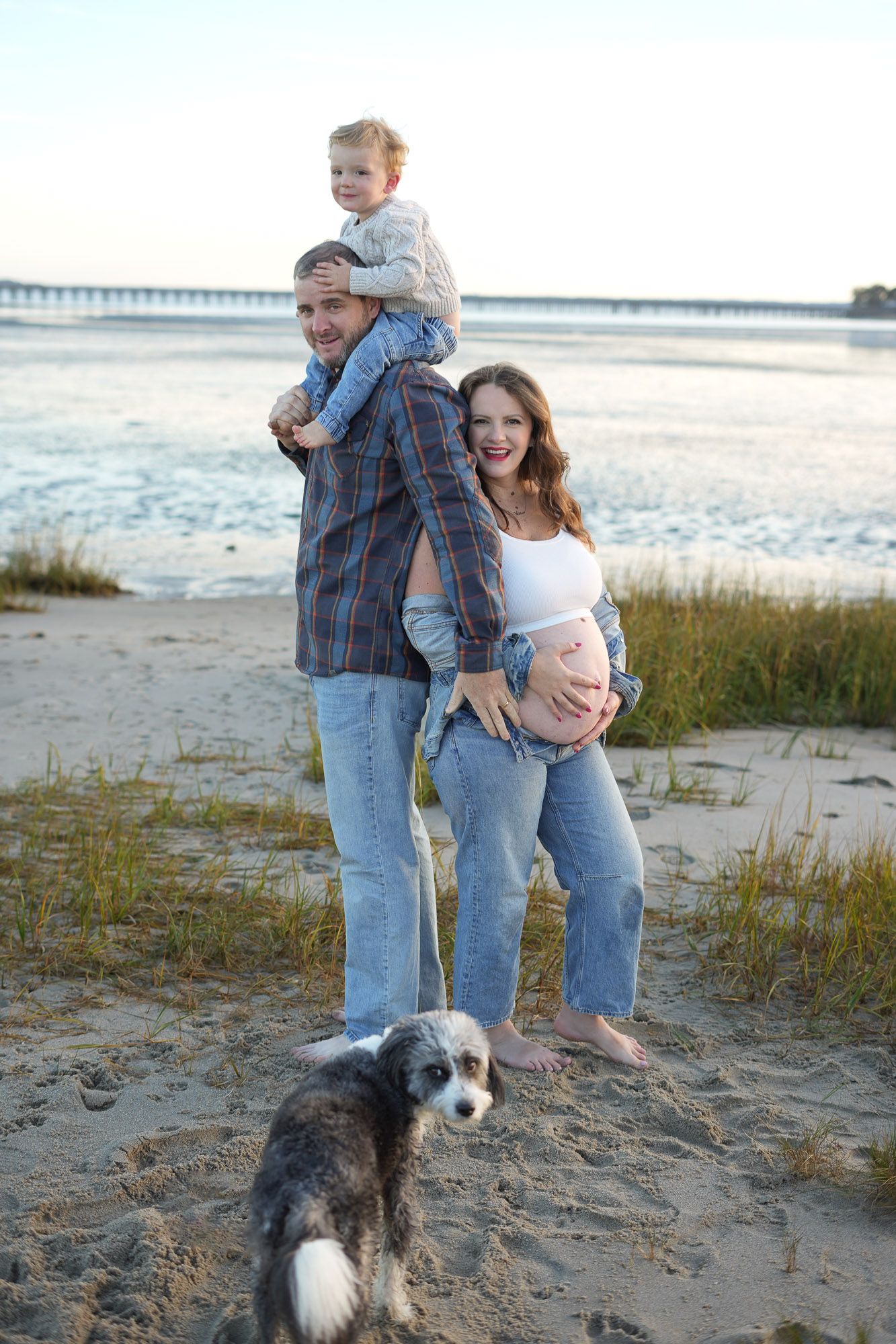 A pregnant family with a toddler on dads shoulders and their dog poses happily on a beach at sunset.