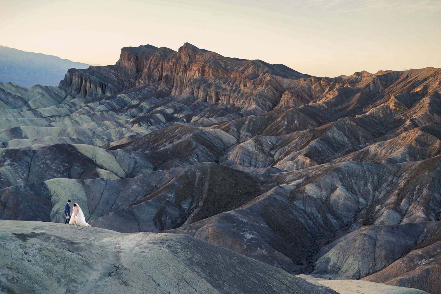Bride and groom tiny figures embracing against vast dramatic Death Valley badlands landscape at dusk.