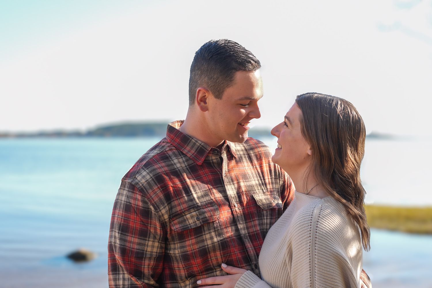 Engaged couple sharing romantic moment near calm waterfront on sunny day.