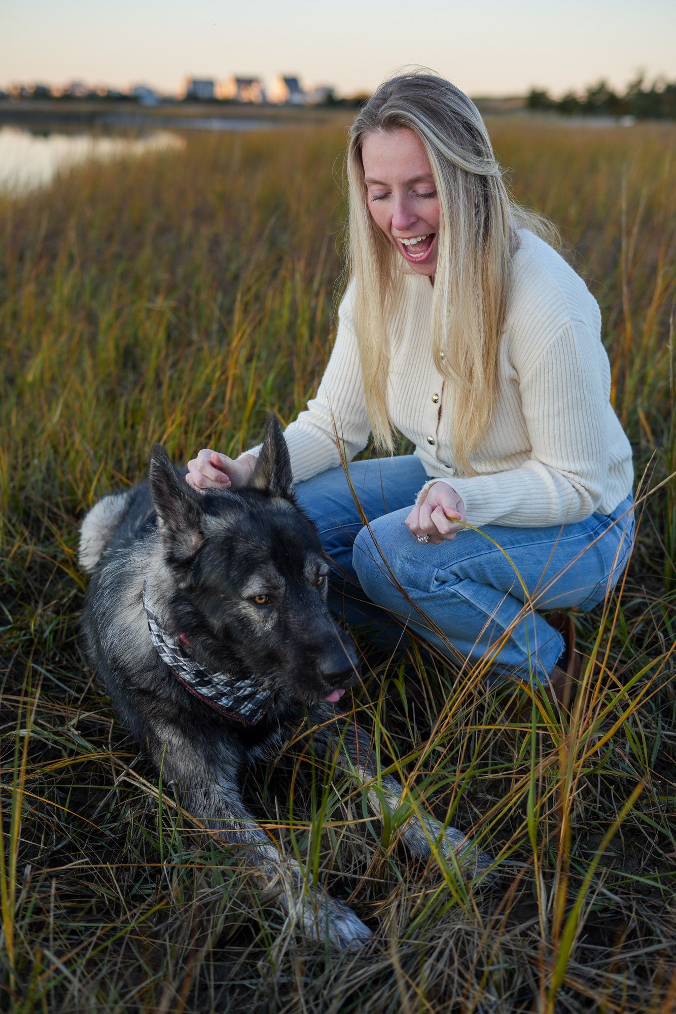 A laughing blonde woman petting her German Shepherd dog in a golden marsh field.