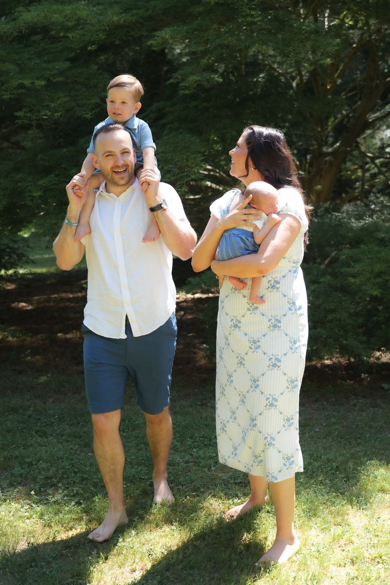 Joyful family of four outdoors, dad carrying toddler on shoulders, mom holding newborn.