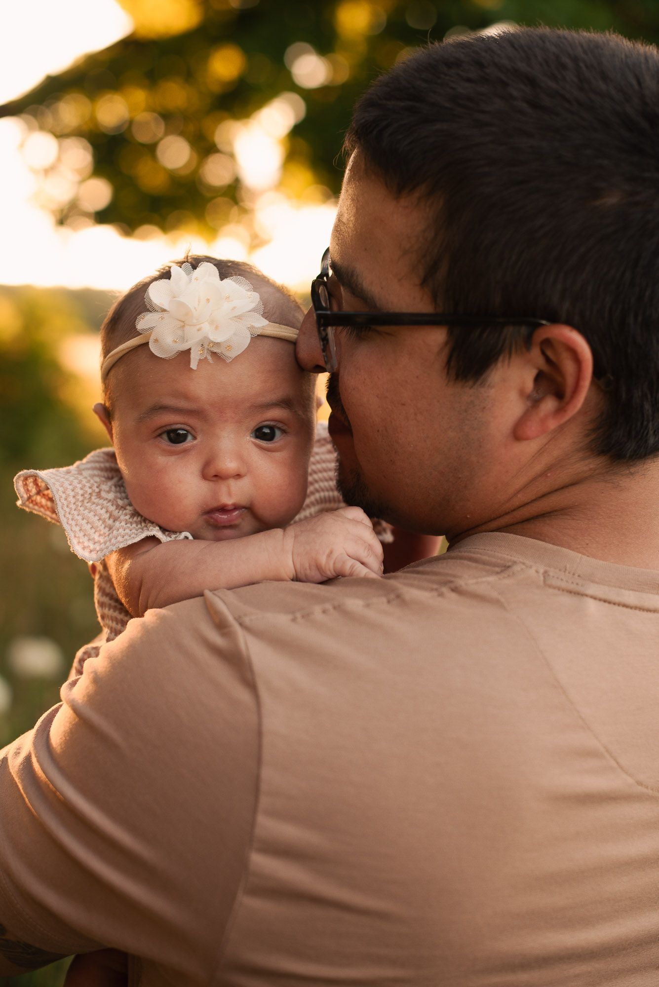 A father holds his newborn baby with a white flower headband close to his cheek outdoors.