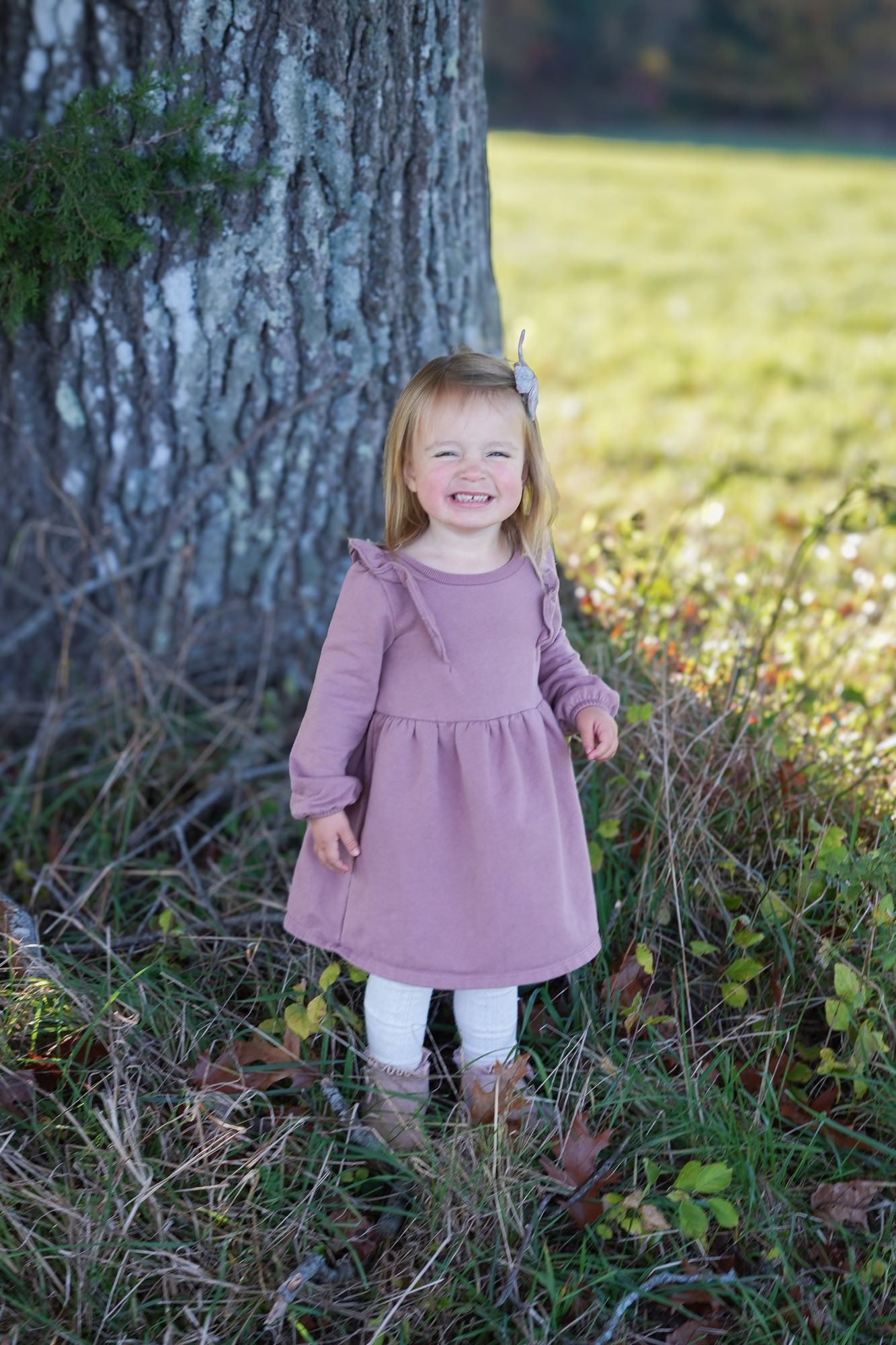Happy toddler girl in pink ruffle dress laughing while standing next to a large tree outdoors.