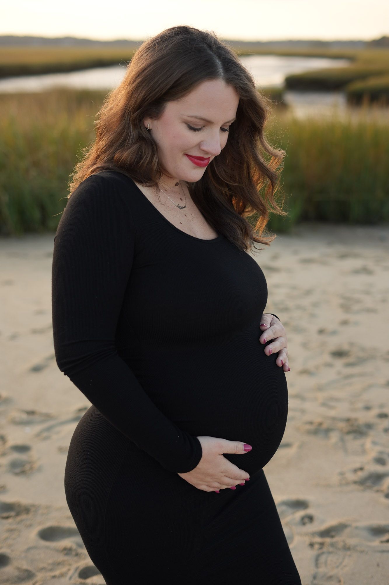 A pregnant woman in a black dress cradles her baby bump while standing on a sandy beach at golden hour.