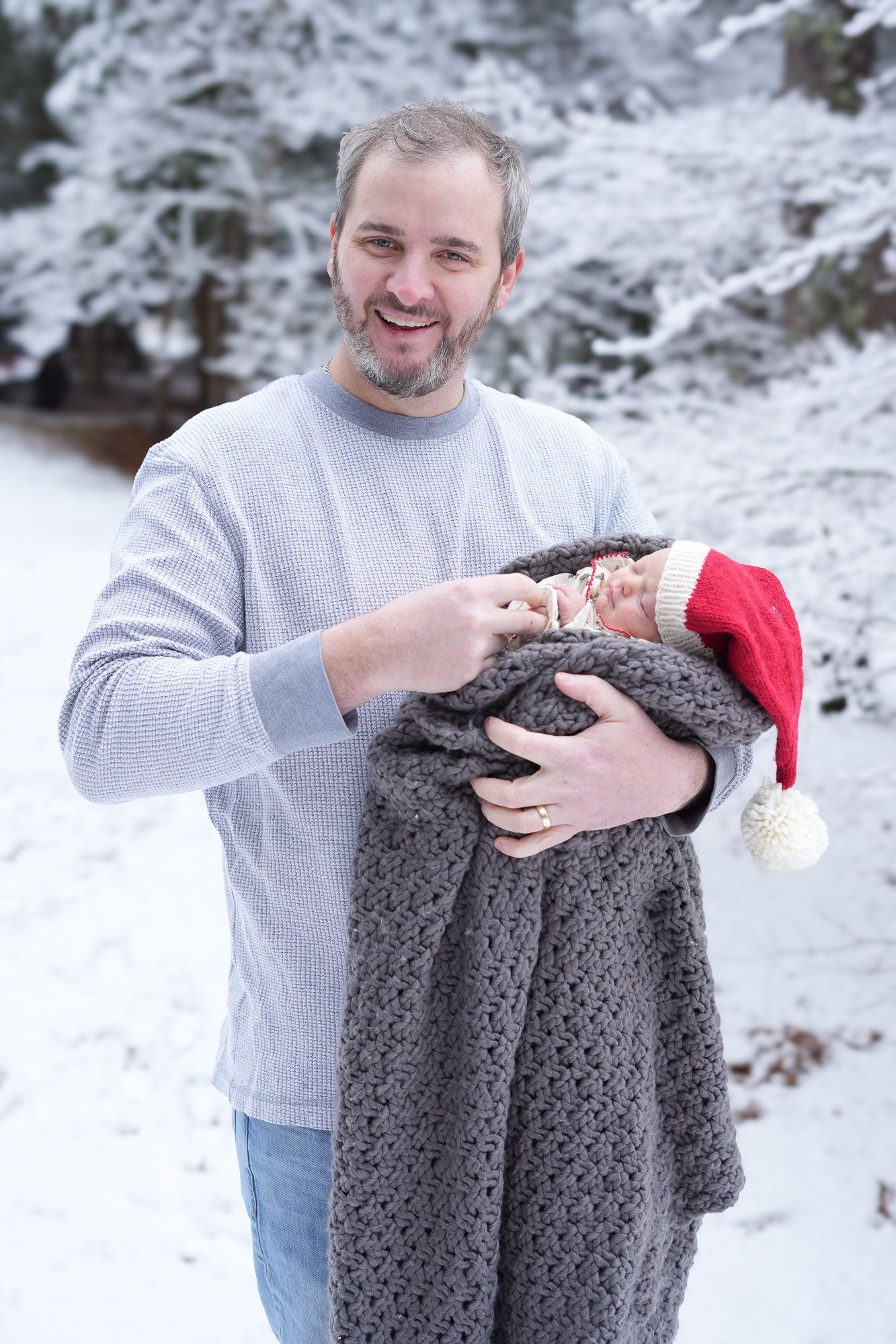 Smiling father pointing at sleeping newborn in gray knit wrap with red Santa hat outdoors in snow.