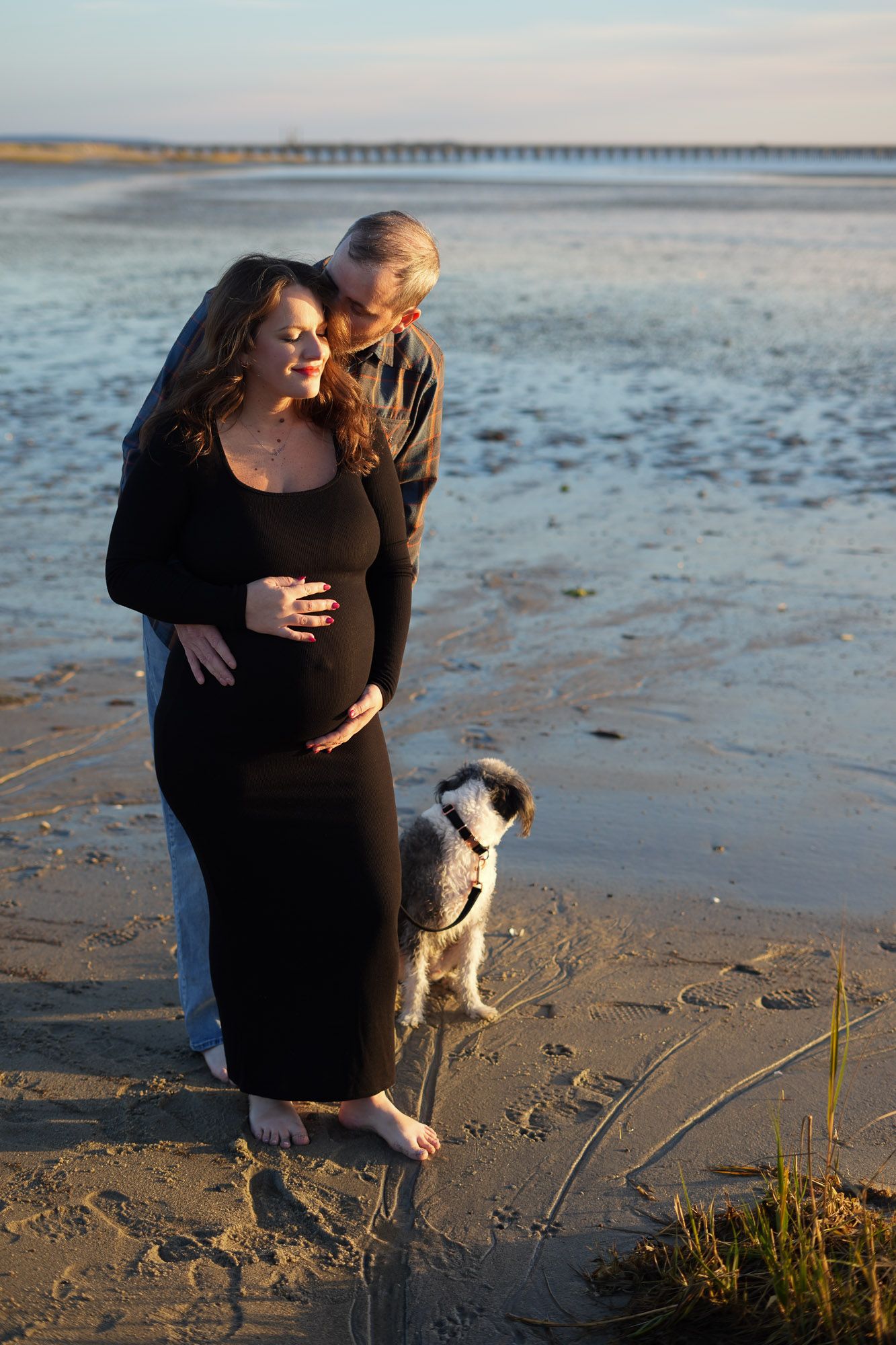 Couple with small dog embracing on beach at sunset, woman in black maternity dress.