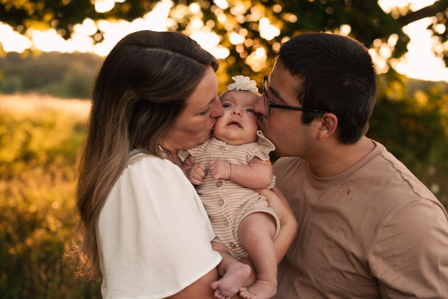 Smiling parents simultaneously kiss their giggling baby on the cheeks in a warm sunset setting.