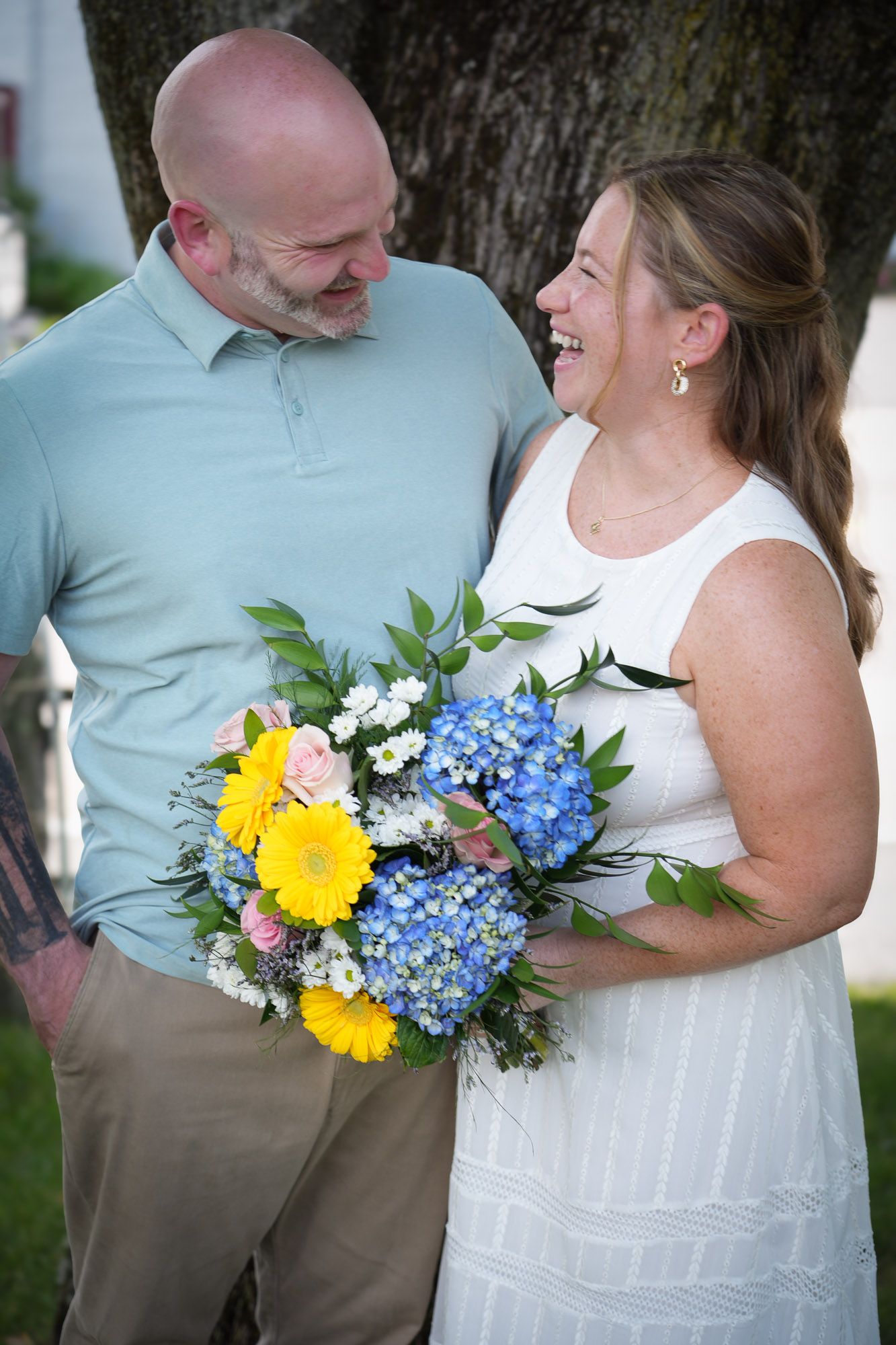 Happy couple sharing a laugh outdoors, bride holding vibrant bouquet of blue hydrangeas and yellow daisies.