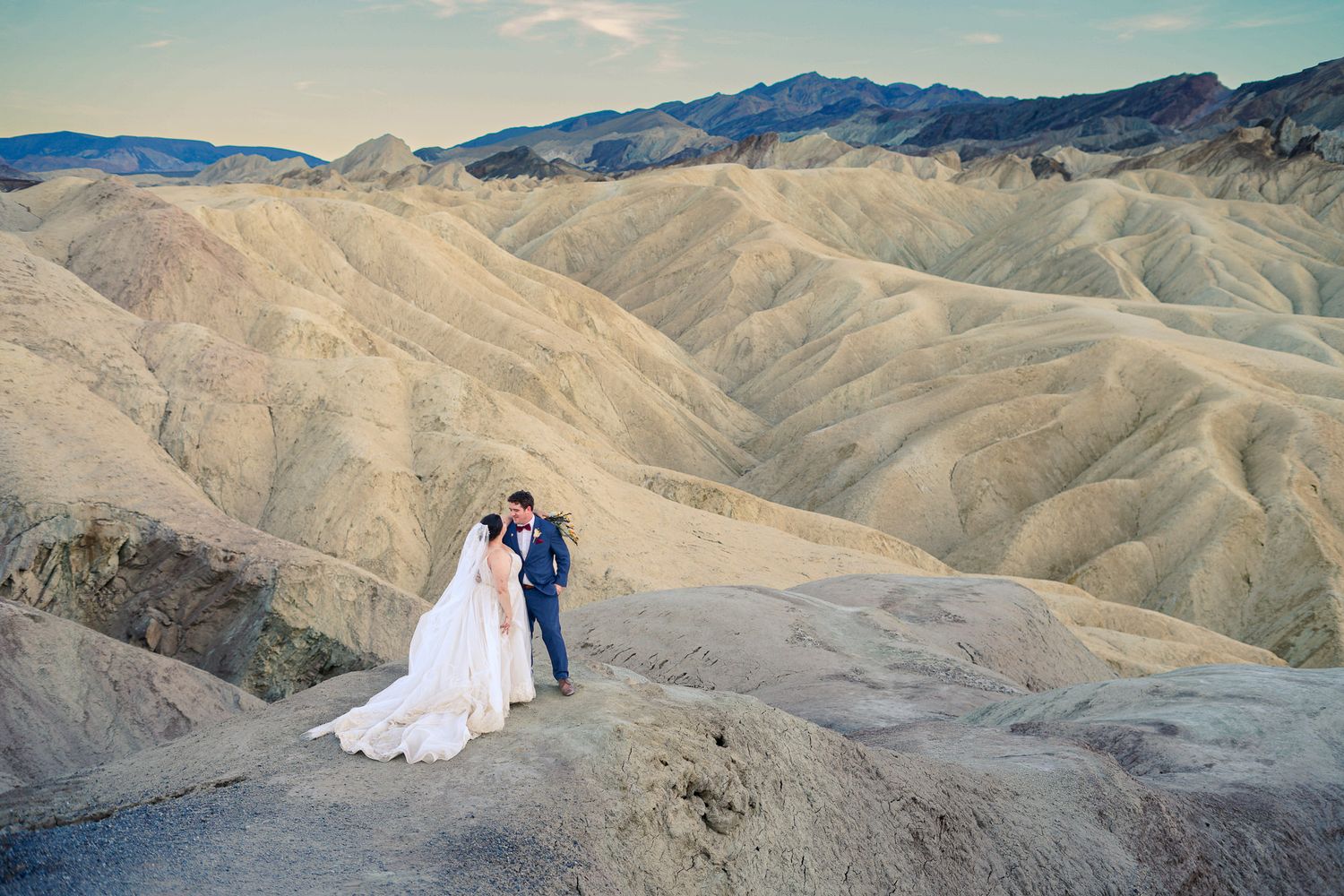 Couple in wedding attire share an intimate moment on rocky overlook with sweeping Death Valley mountain views behind them.