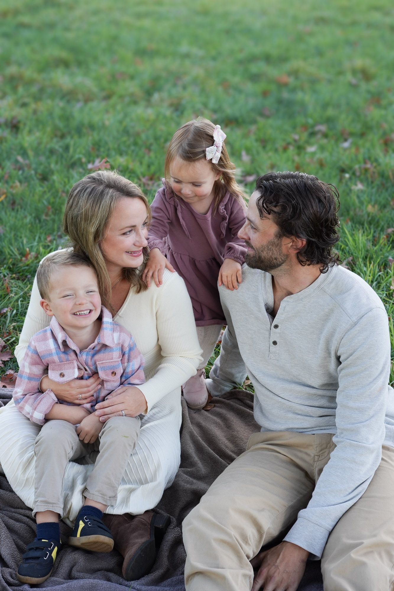 Family of four sitting together on a blanket in a grassy park, smiling and laughing together.