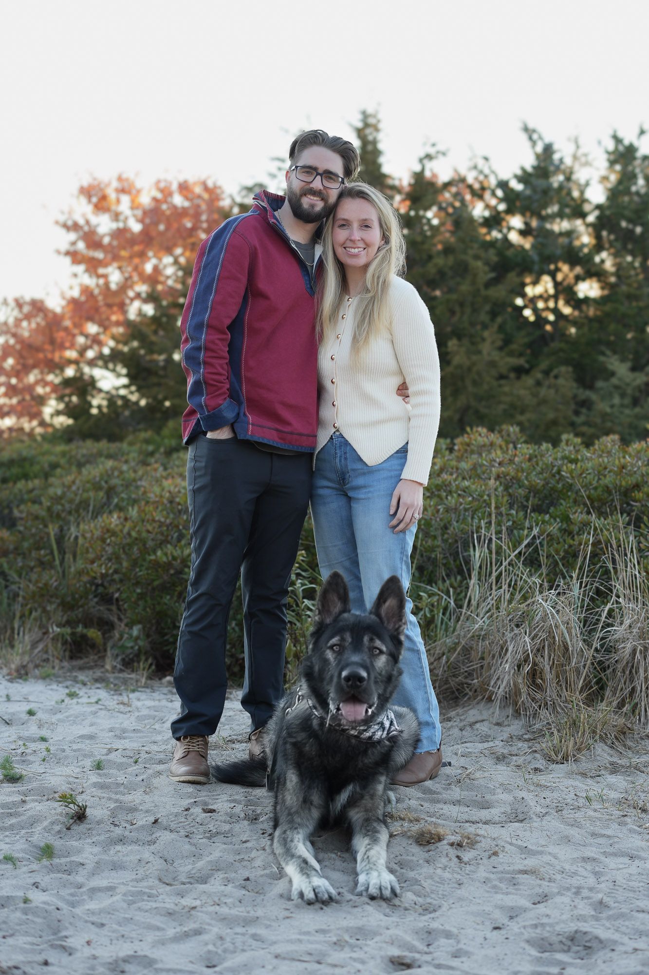 Happy couple standing with their German Shepherd dog on a sandy beach surrounded by autumn foliage.