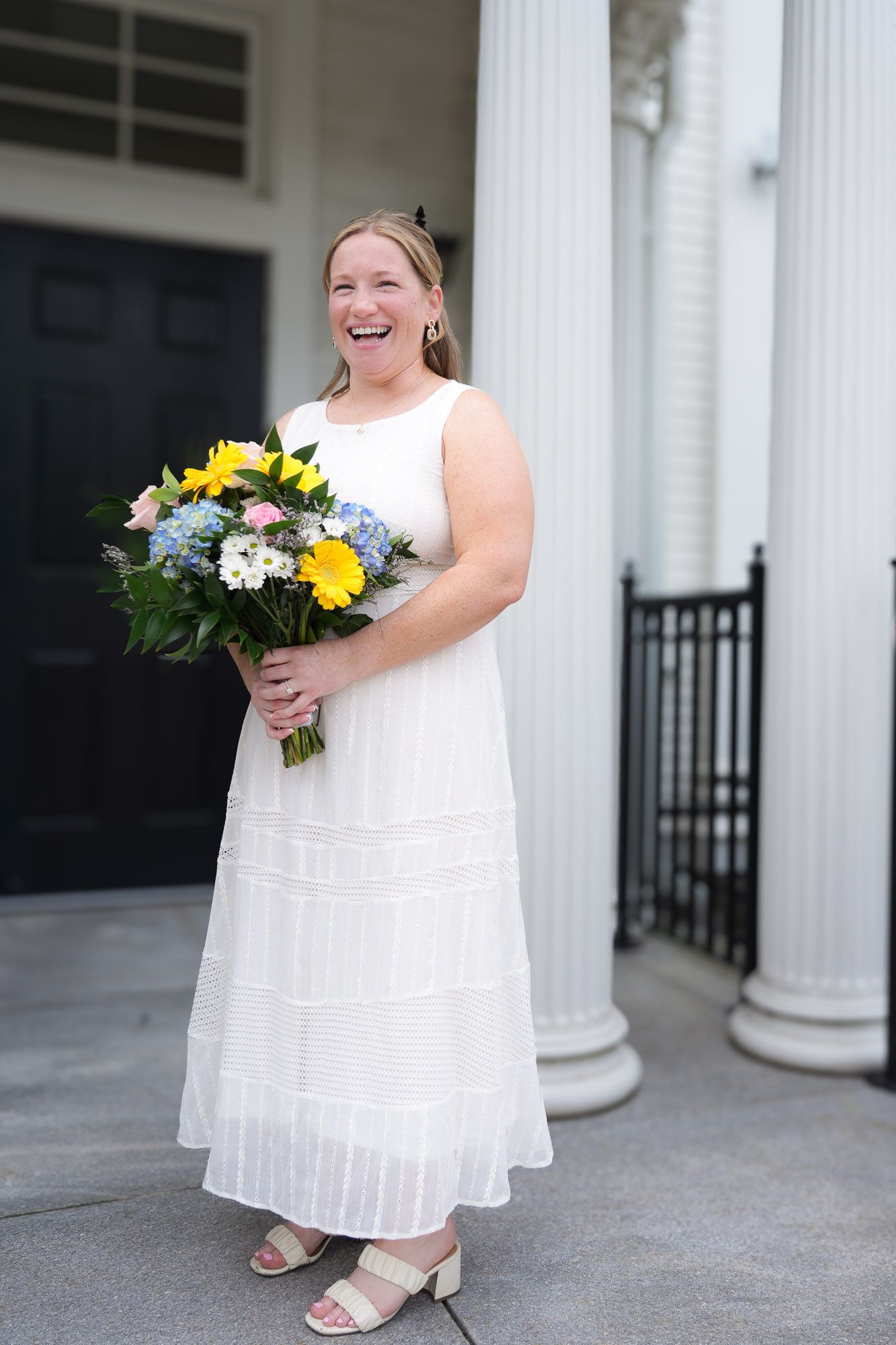A bride in a white dress laughs joyfully while holding a colorful bouquet outside a building with columns.