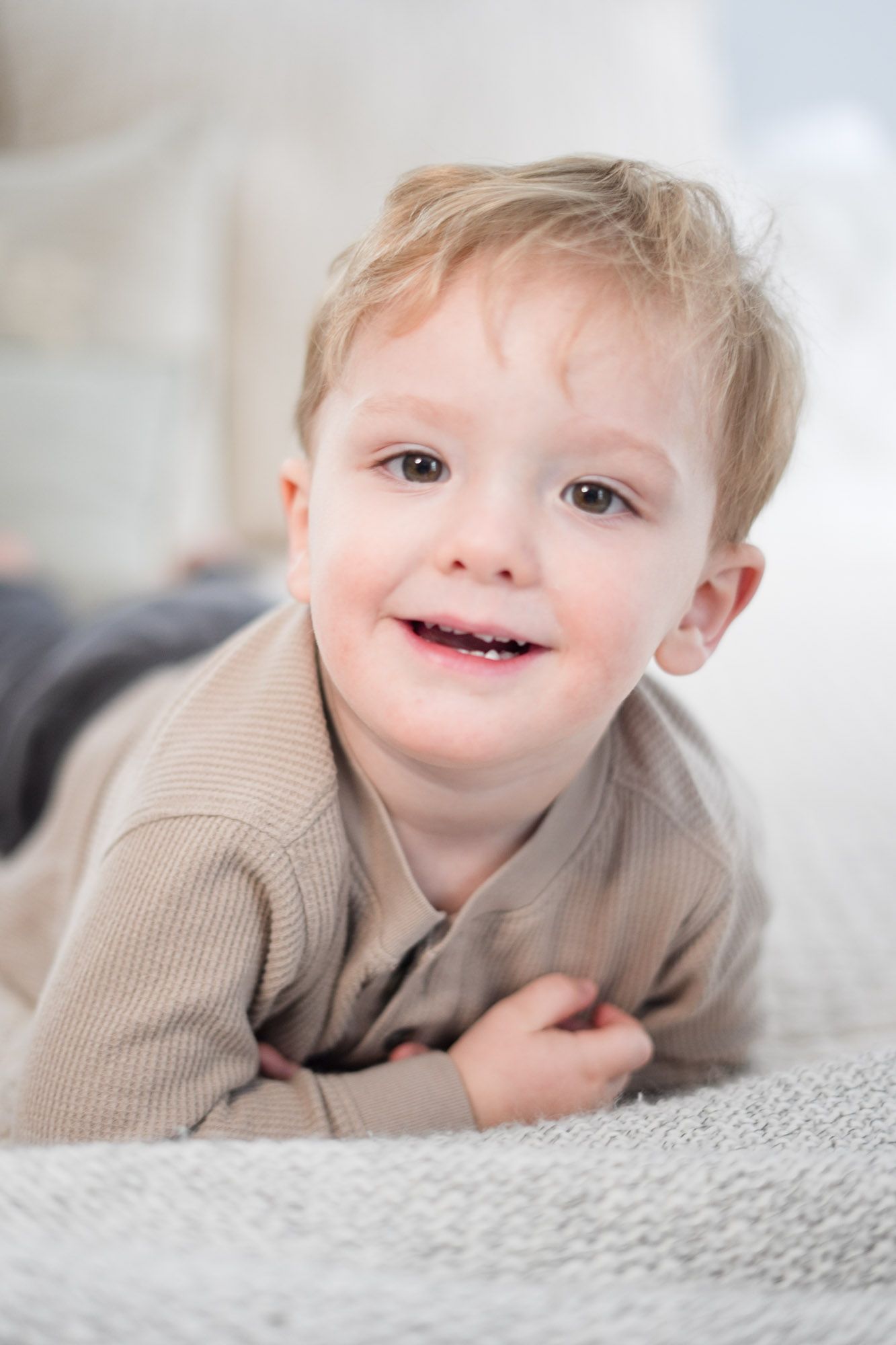 Adorable smiling toddler with blonde hair lying on a white knit blanket looking at the camera.