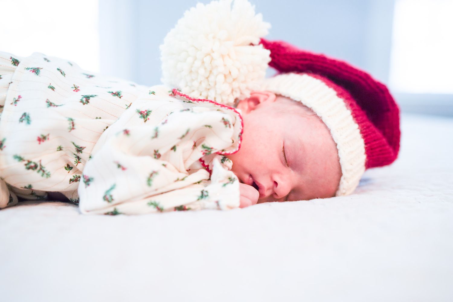 Sleeping newborn baby in floral onesie wearing red and white Santa Claus hat on white blanket.
