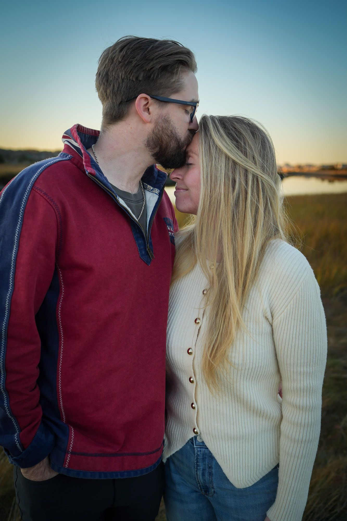Man gently kissing womans forehead in a grassy marsh field at dusk.