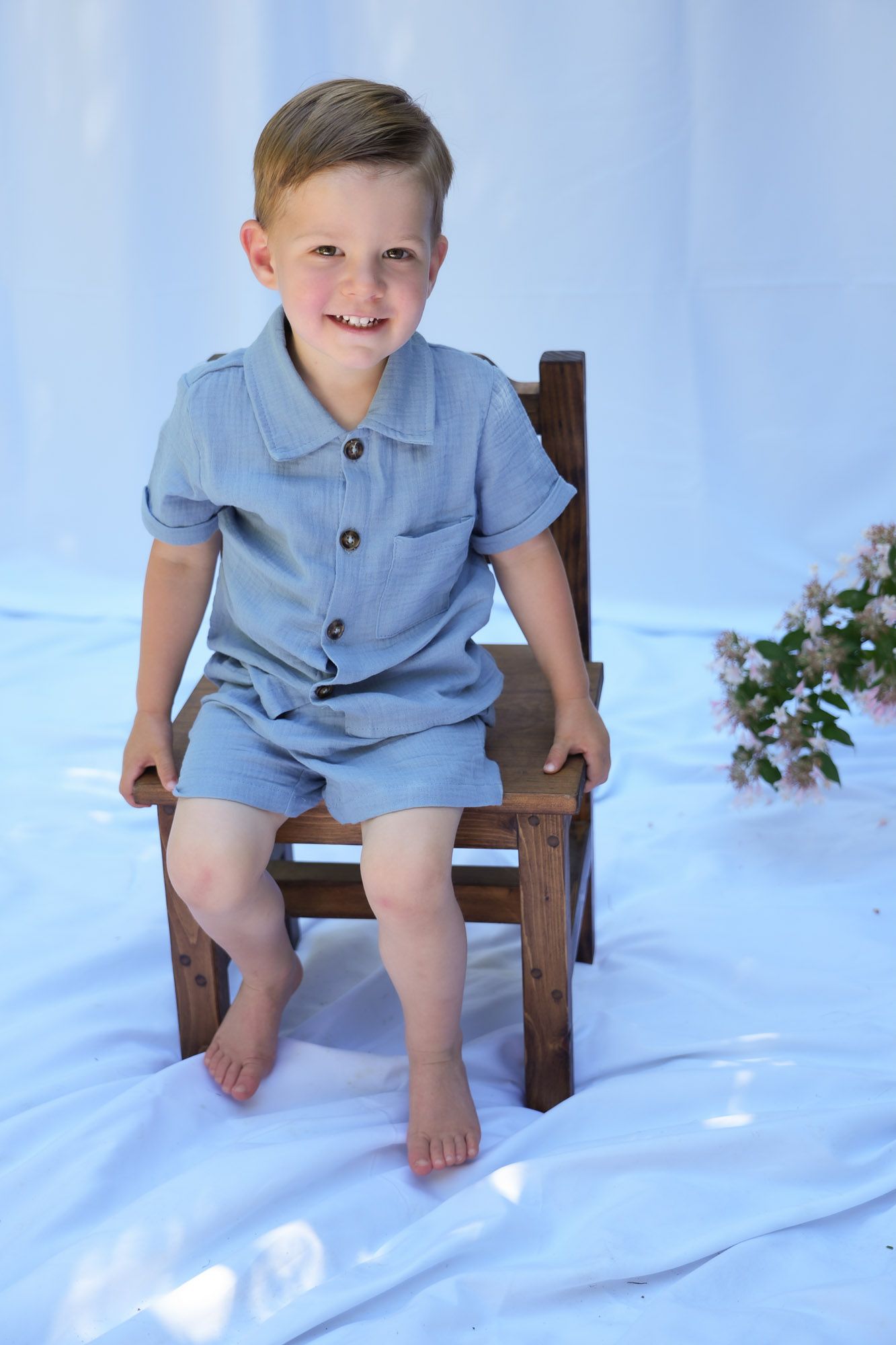 Happy young boy in blue linen outfit sitting on wooden chair smiling at camera.