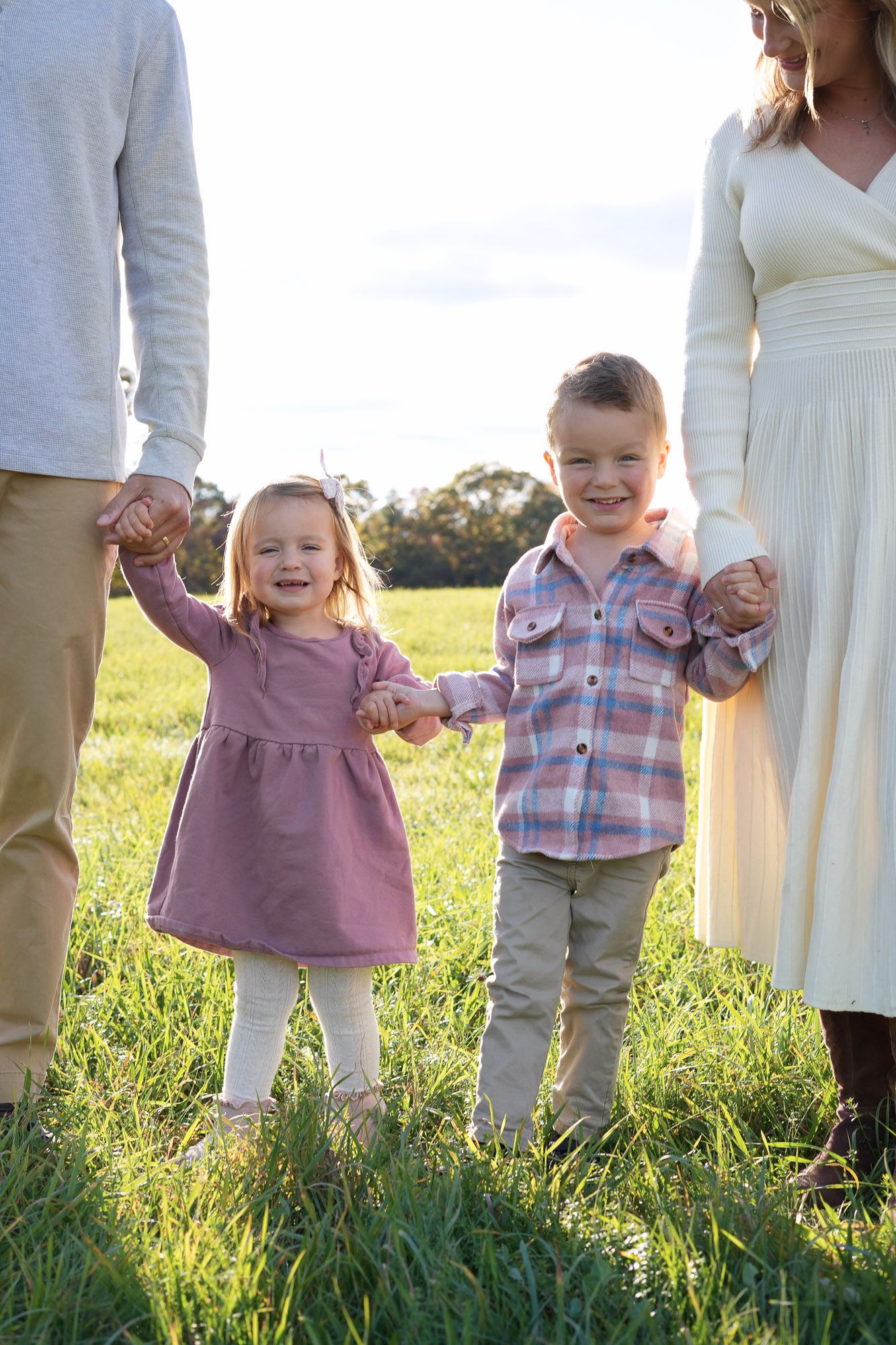 Two young children holding parents hands in a sunny green field.