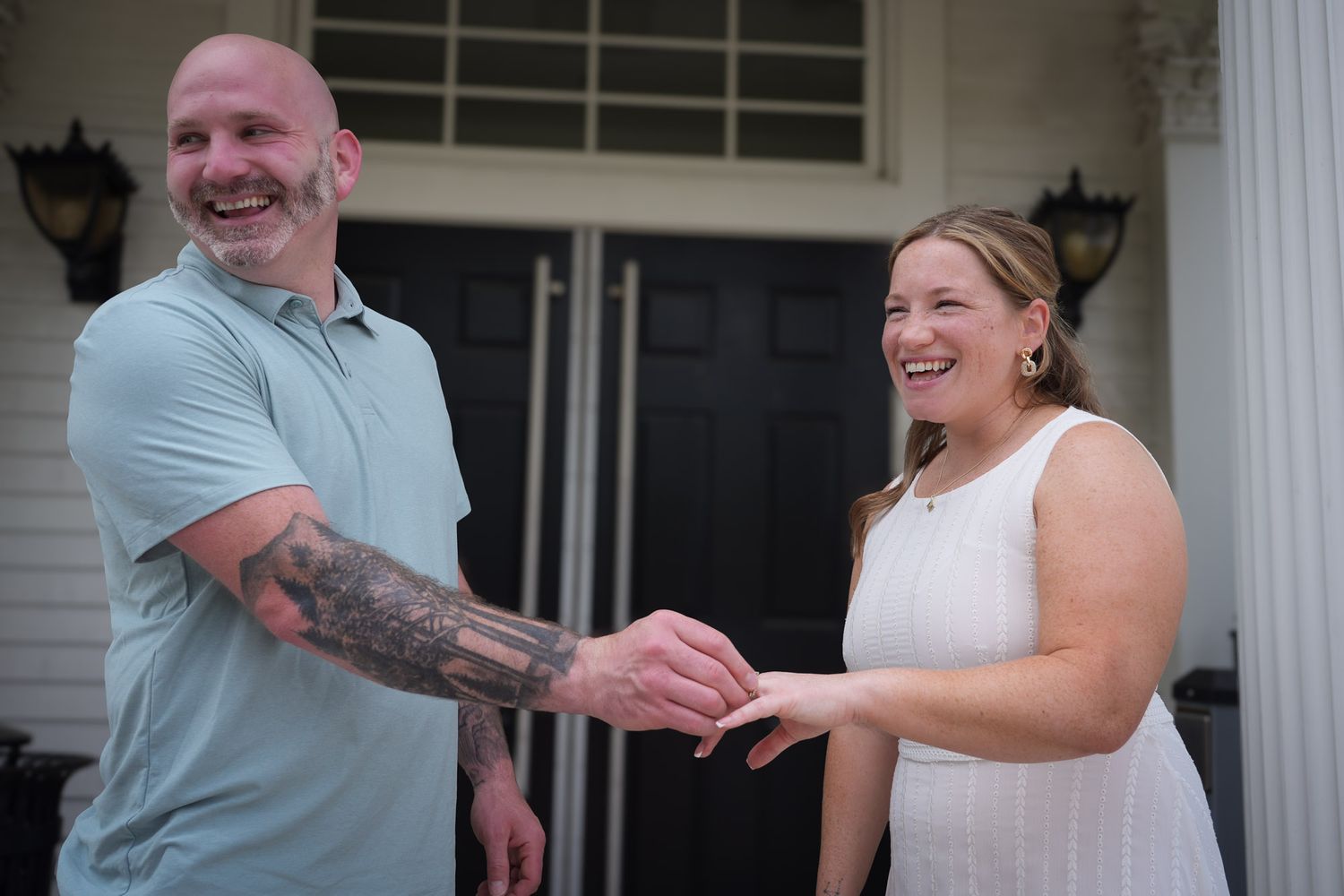 Smiling couple sharing a lighthearted moment during ring exchange ceremony on white columned front porch.