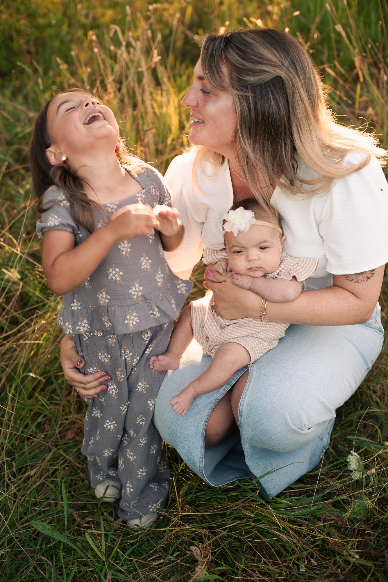 Mother laughing with young daughter and newborn baby in sunny grass field.