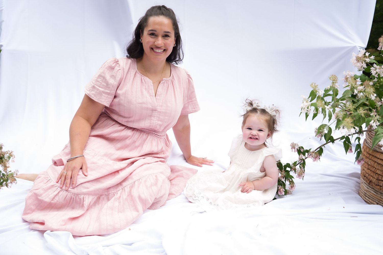 Smiling mother in pink dress sitting with baby girl in white dress during outdoor photo shoot.