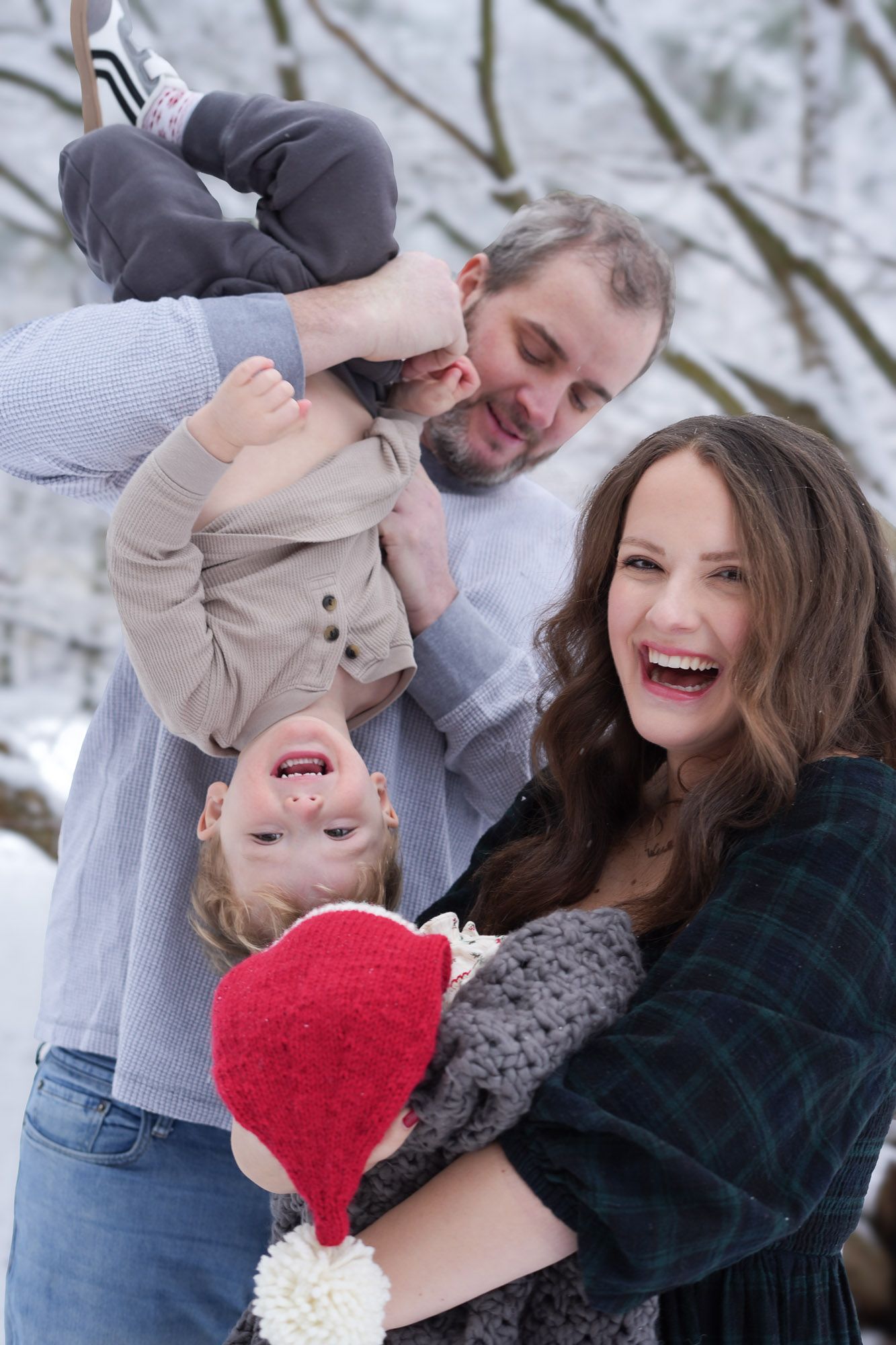 Joyful family laughing in snowy winter setting with father holding toddler upside down and mother with newborn.