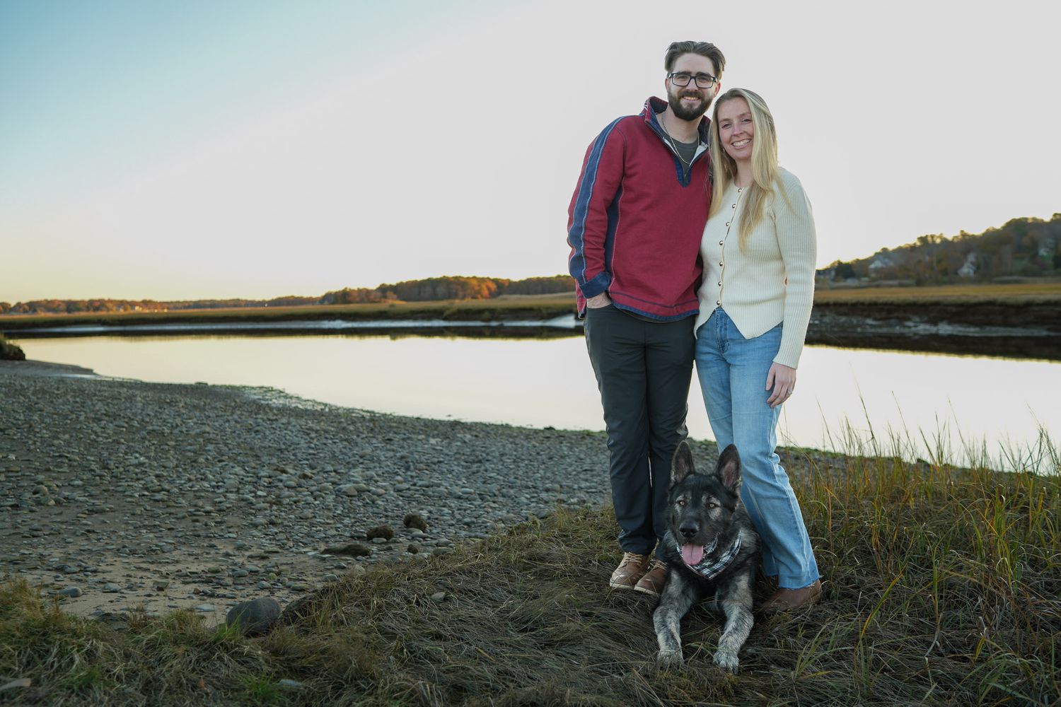Couple smiling with their German Shepherd dog by a scenic marsh during sunset.
