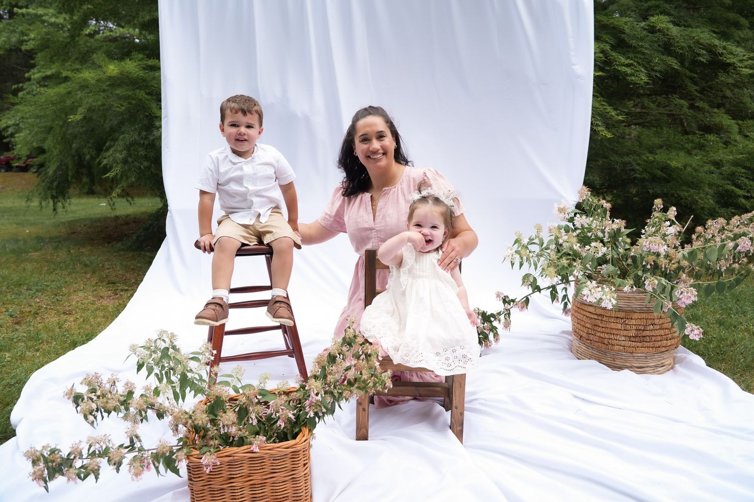 Mother in pink dress smiling with two young children amid floral arrangements at outdoor photo shoot.
