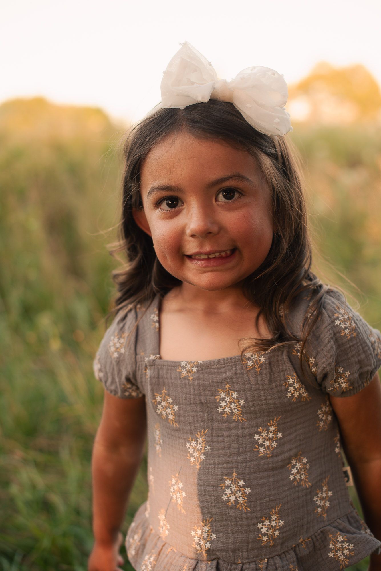 A cute little girl in a floral dress and white bow headband smiles sweetly in a sunlit outdoor field.