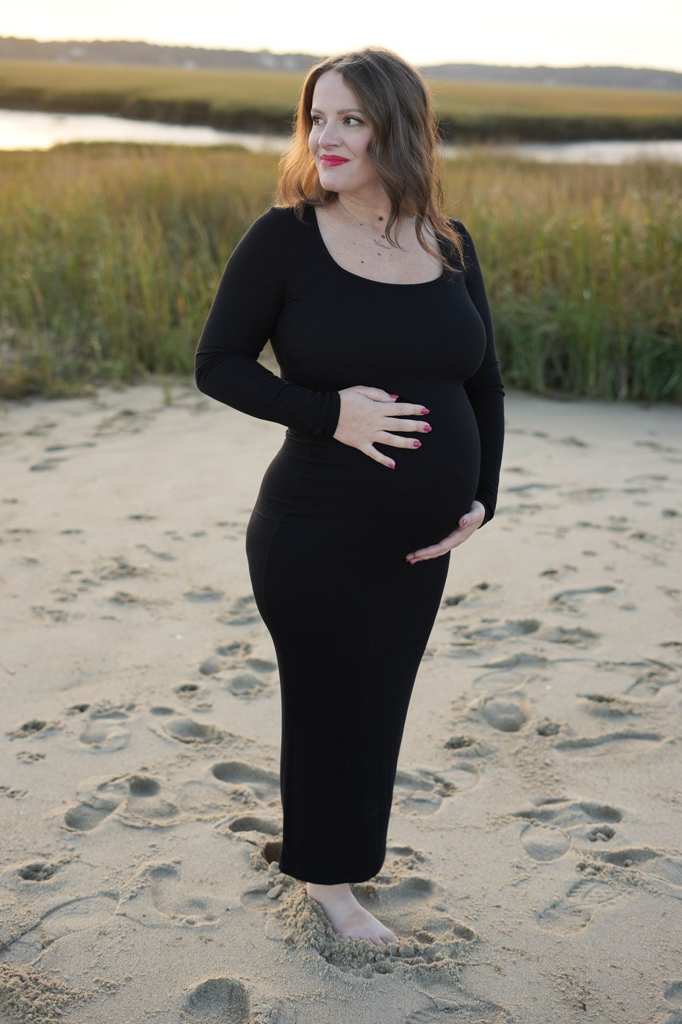 Smiling pregnant woman in black long-sleeve dress cradling bump on beach at dusk.