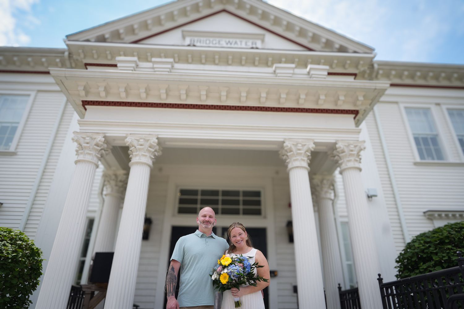 Smiling newlyweds standing before a white columned Bridgewater building with floral bouquet.