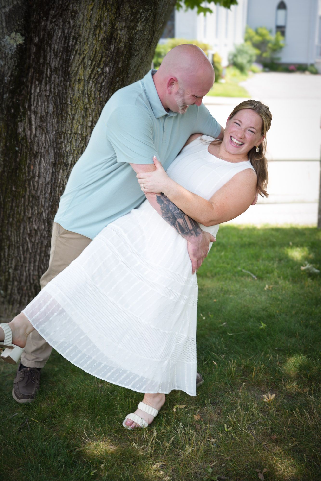A bald man playfully dips a laughing woman in a white dress outdoors near a tree.