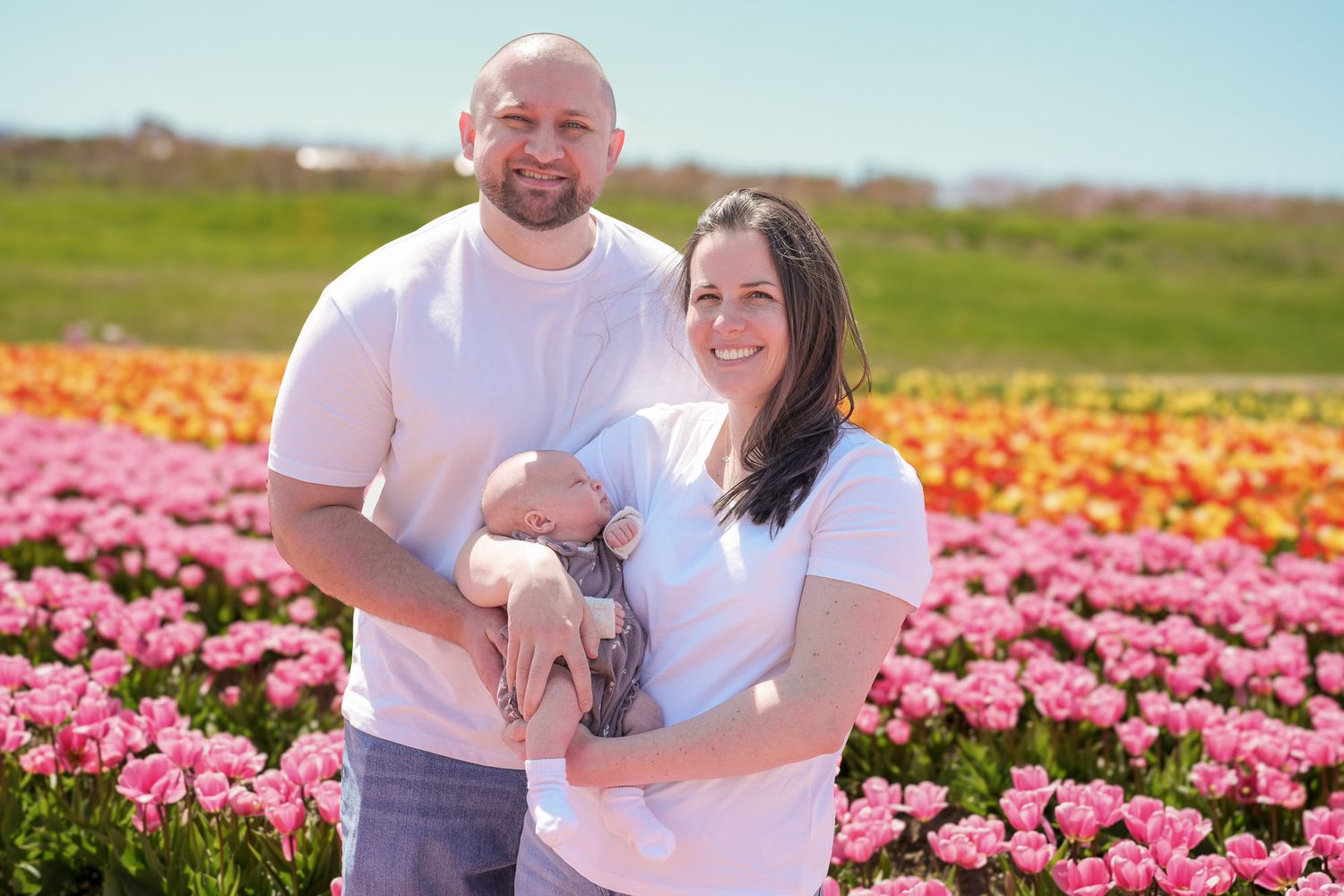 A smiling couple in matching white shirts holds their newborn baby in a vibrant pink tulip field.