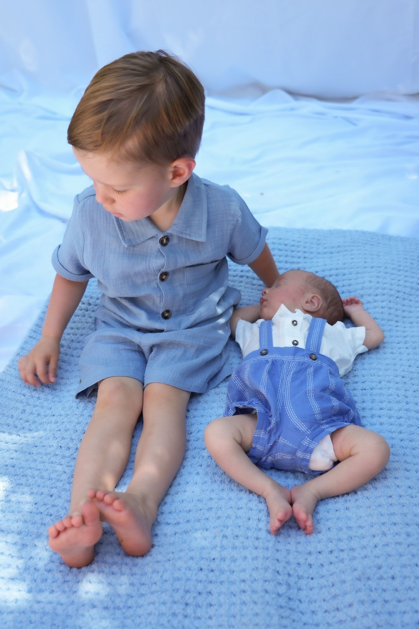 Toddler boy in blue romper sitting beside sleeping newborn sibling on blue knit blanket.
