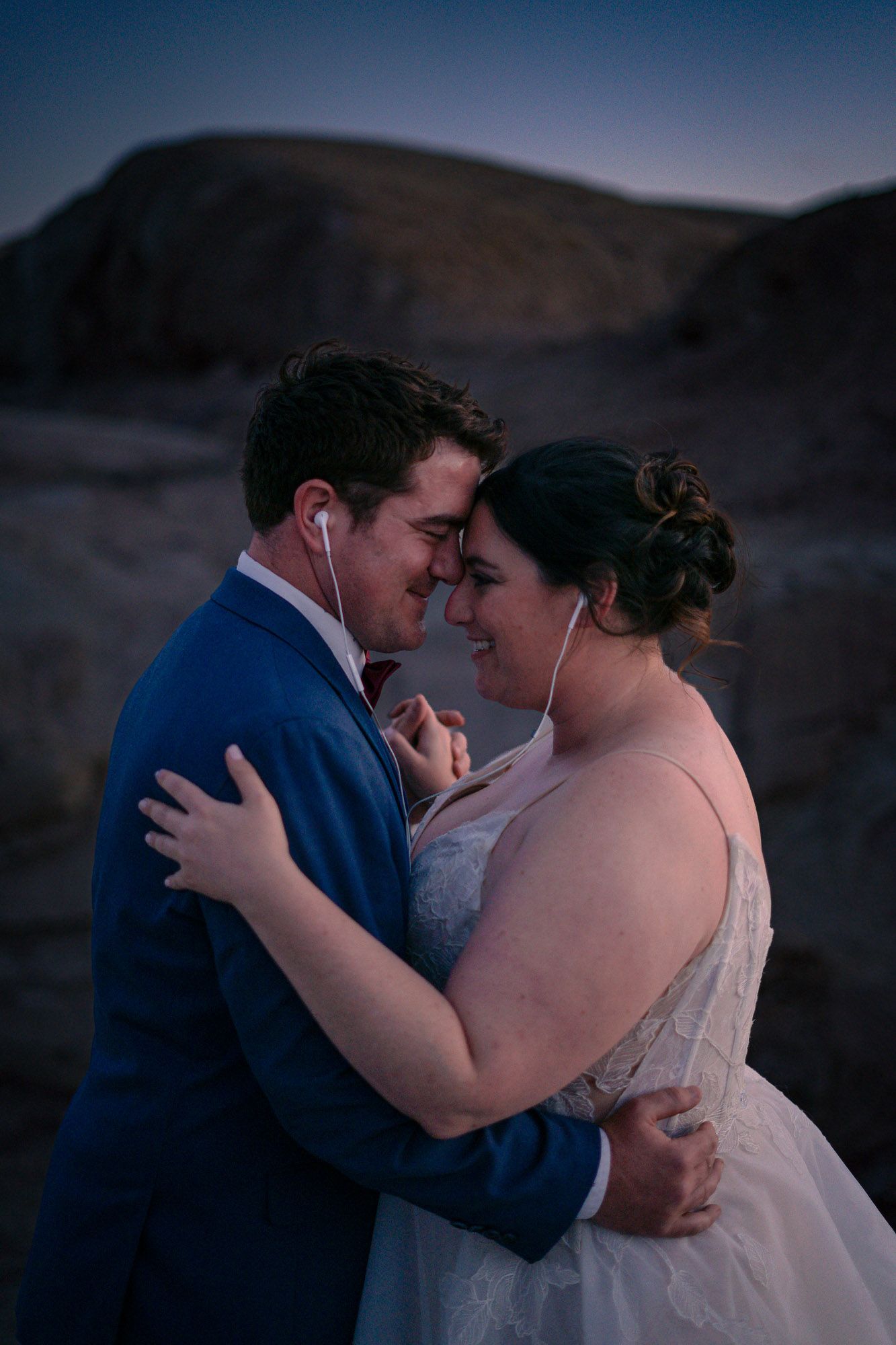Bride and groom share a tender forehead touch while sharing earbuds dancing together at twilight in the desert.