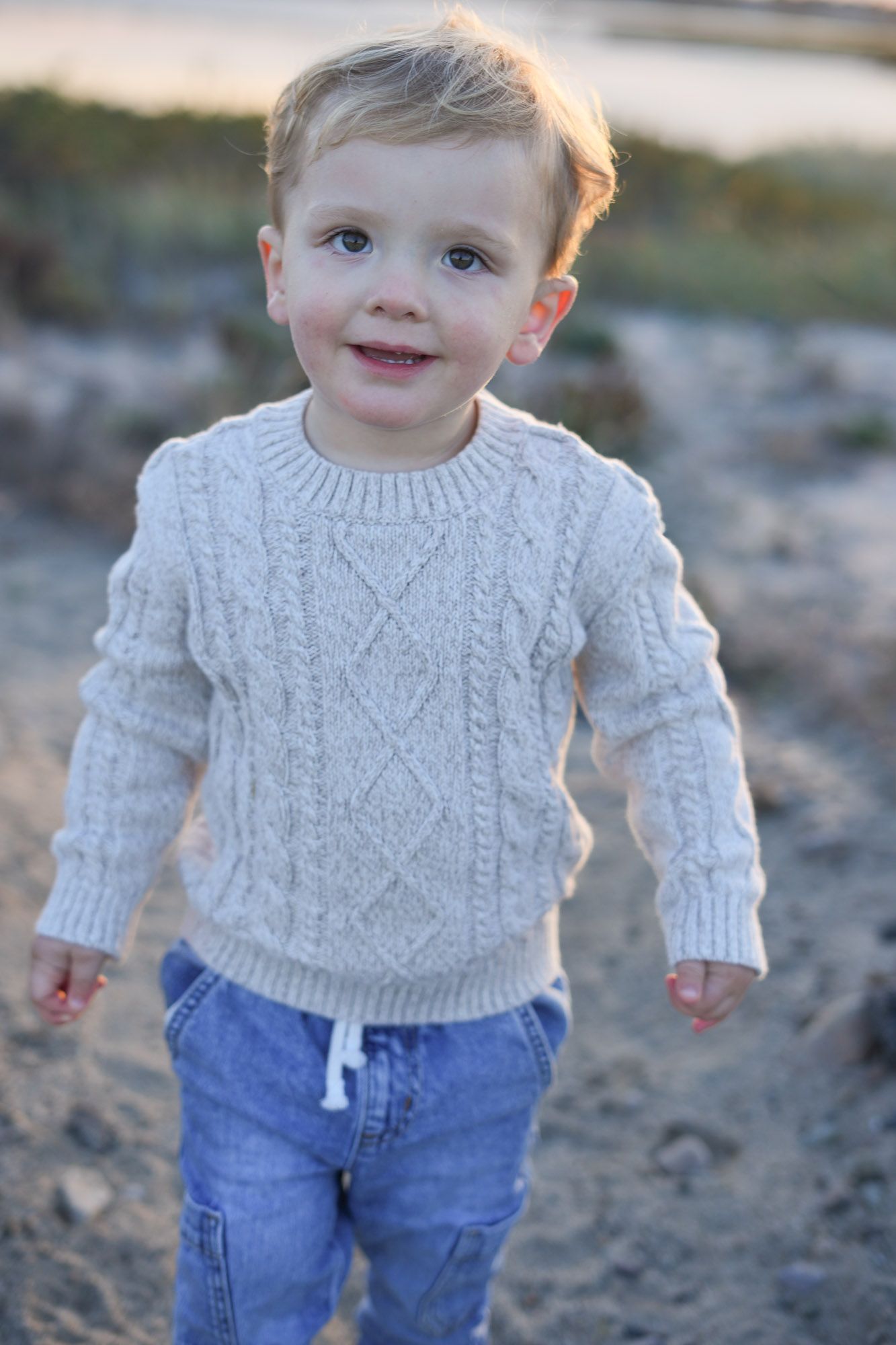 Adorable blond toddler boy in cable knit sweater and jeans smiling on beach.
