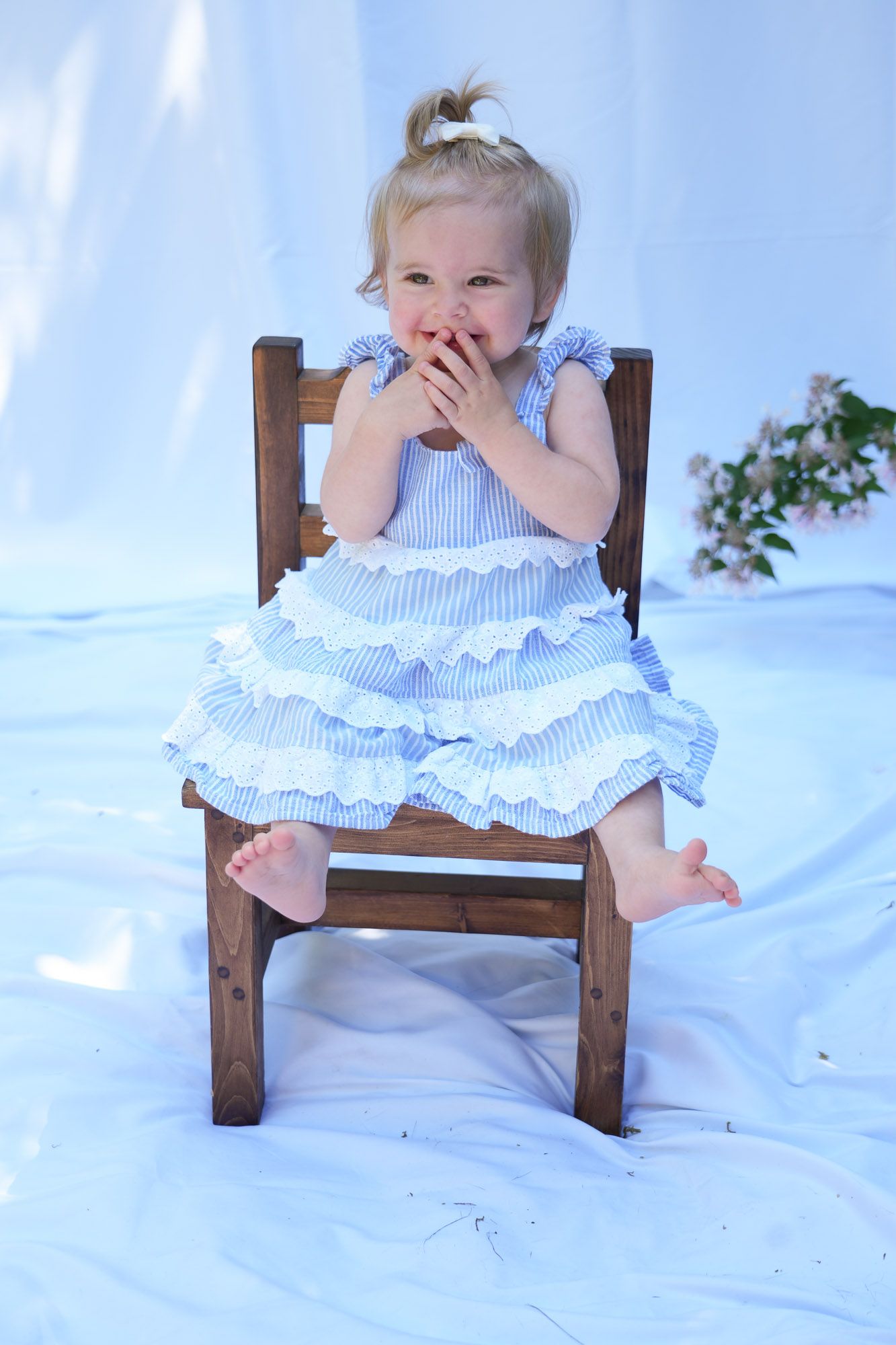 Adorable toddler girl in blue ruffled dress sitting on wooden chair covering her mouth.