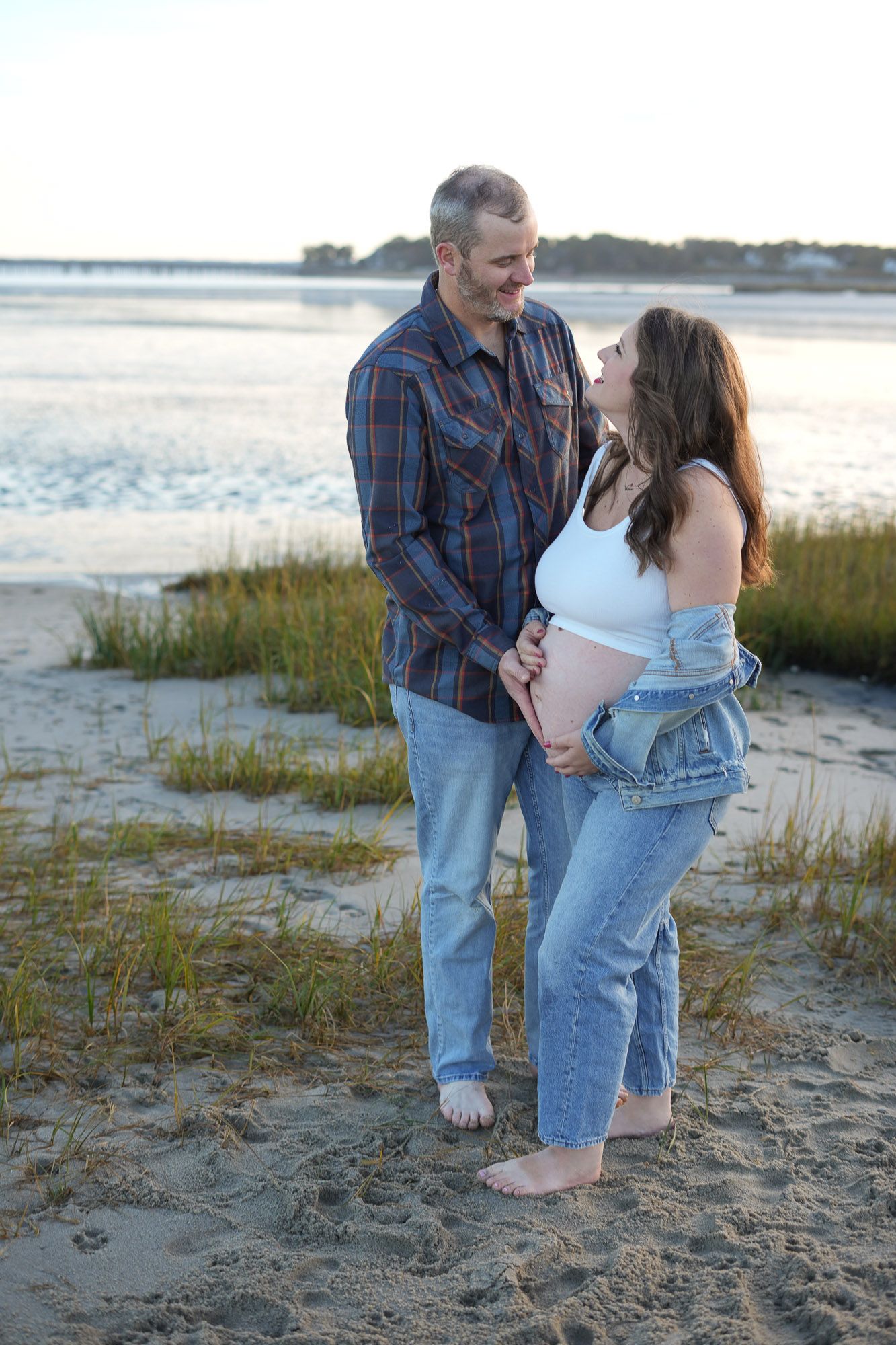 Couple holding pregnant belly together smiling at each other on sandy beach at golden hour.