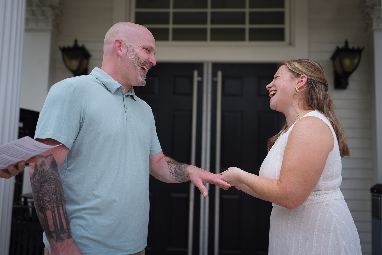 Bride and groom laughing joyfully during outdoor wedding ceremony on white columned porch in daylight.