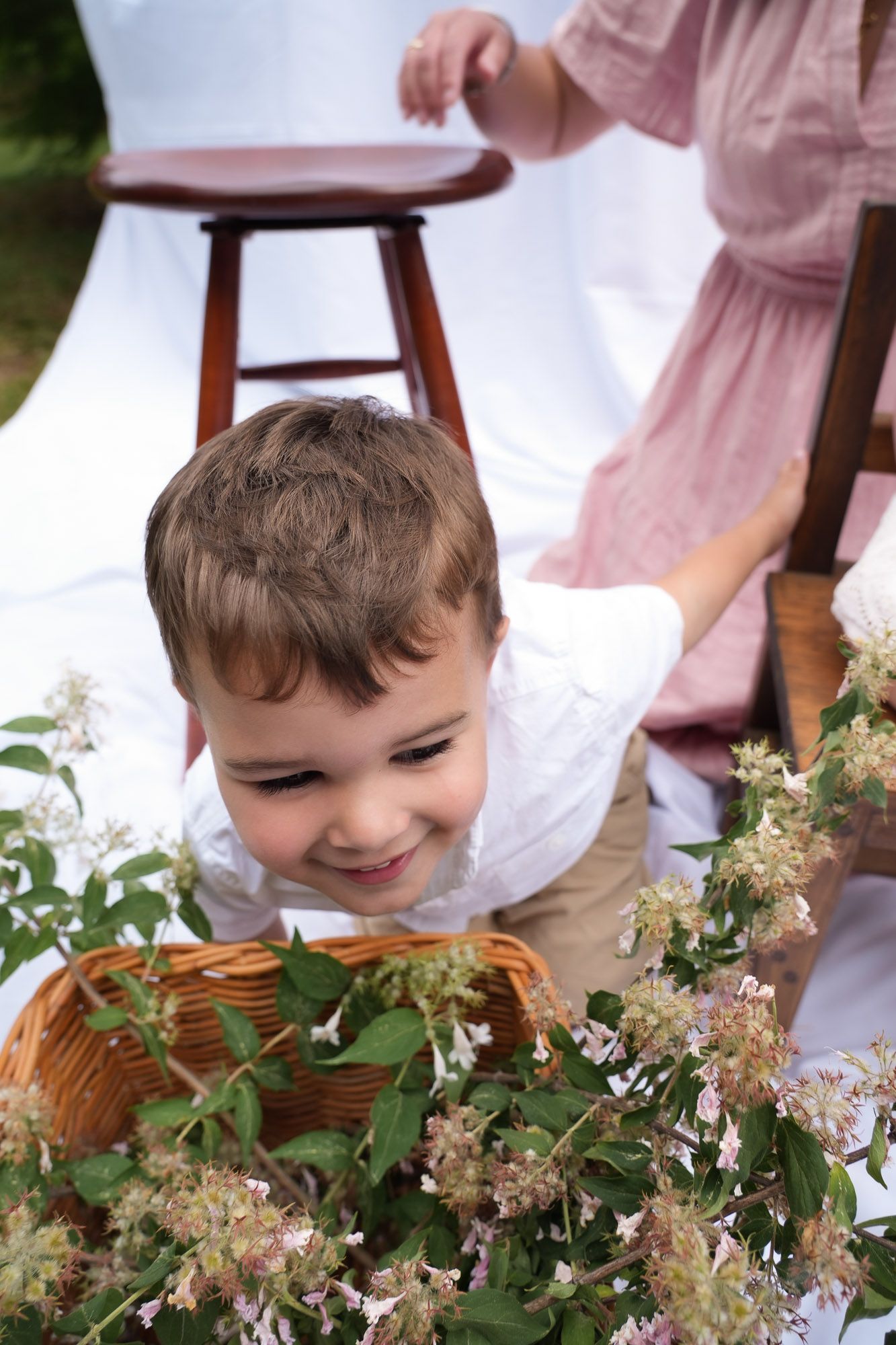 Smiling toddler boy in white shirt peeking over wicker basket of flowers at outdoor photo shoot.