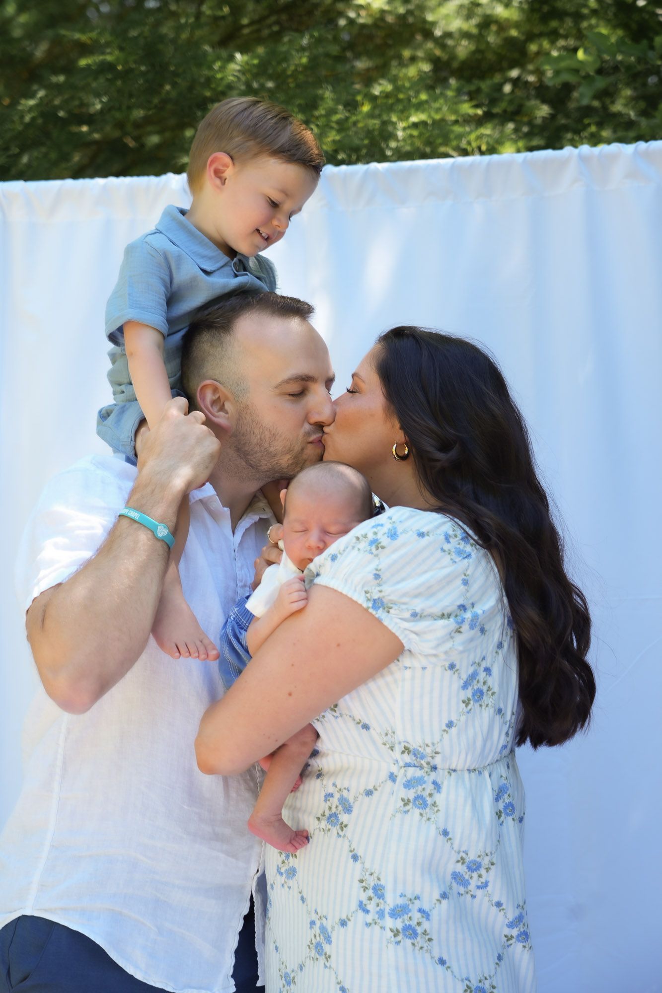Parents kissing while father holds toddler on shoulders and mother holds newborn.
