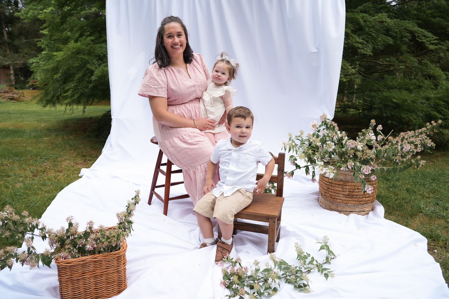 Pregnant woman in pink dress poses outdoors with toddler girl and young boy on wooden chair with flower baskets.