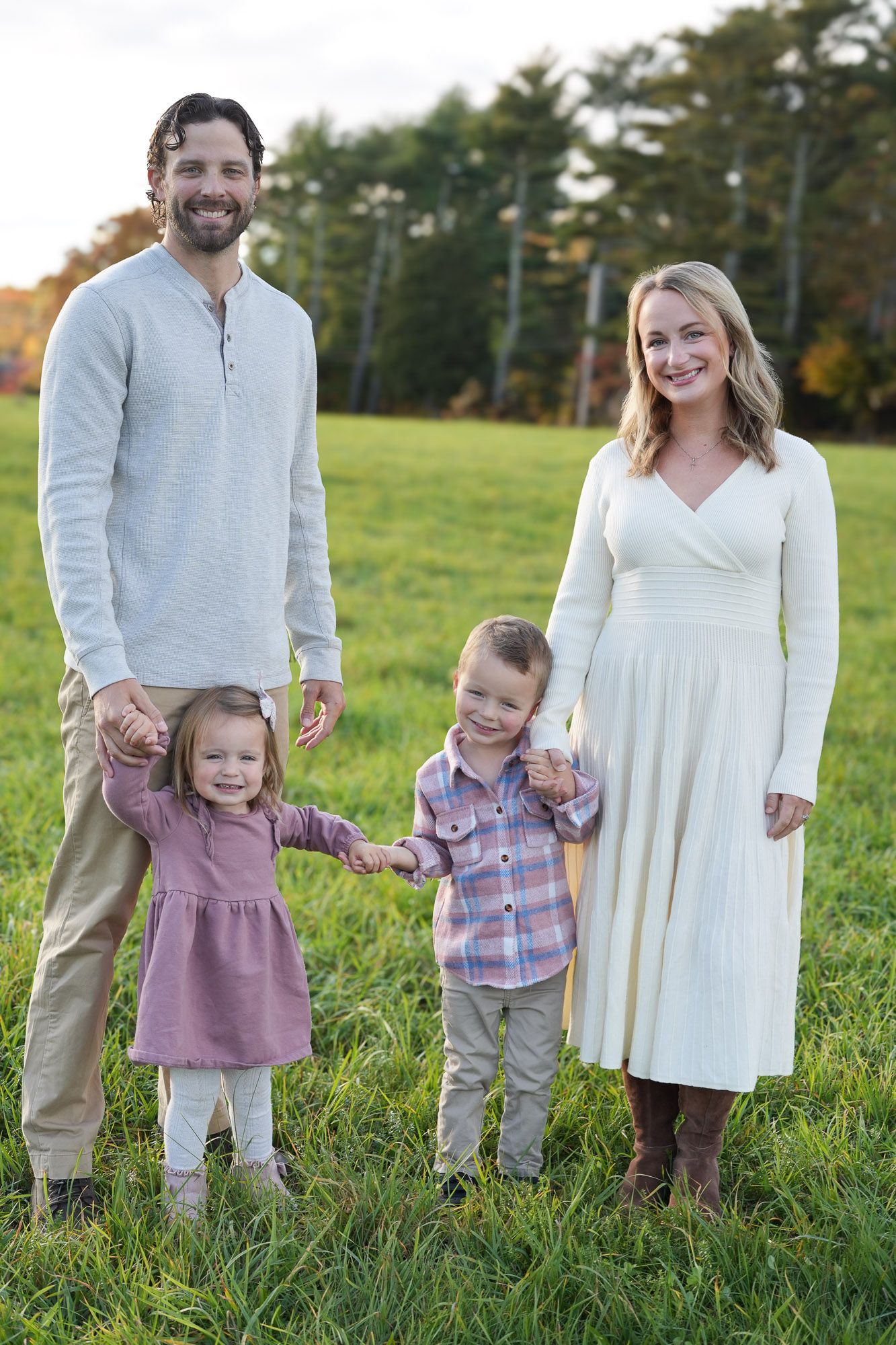 Happy family of four holding hands in a green field at sunset, two young children between smiling parents.