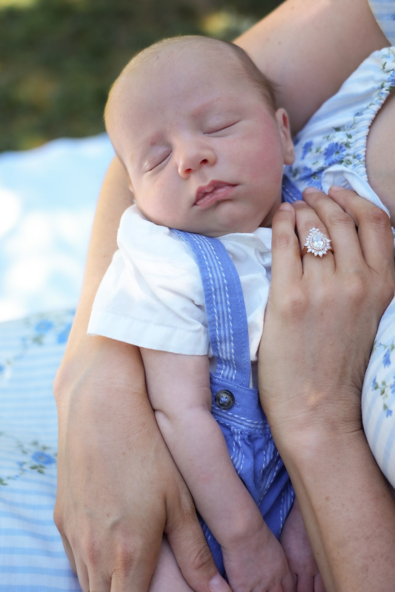 A sleeping newborn in blue suspenders rests peacefully in their mothers arms, showcasing a diamond ring.
